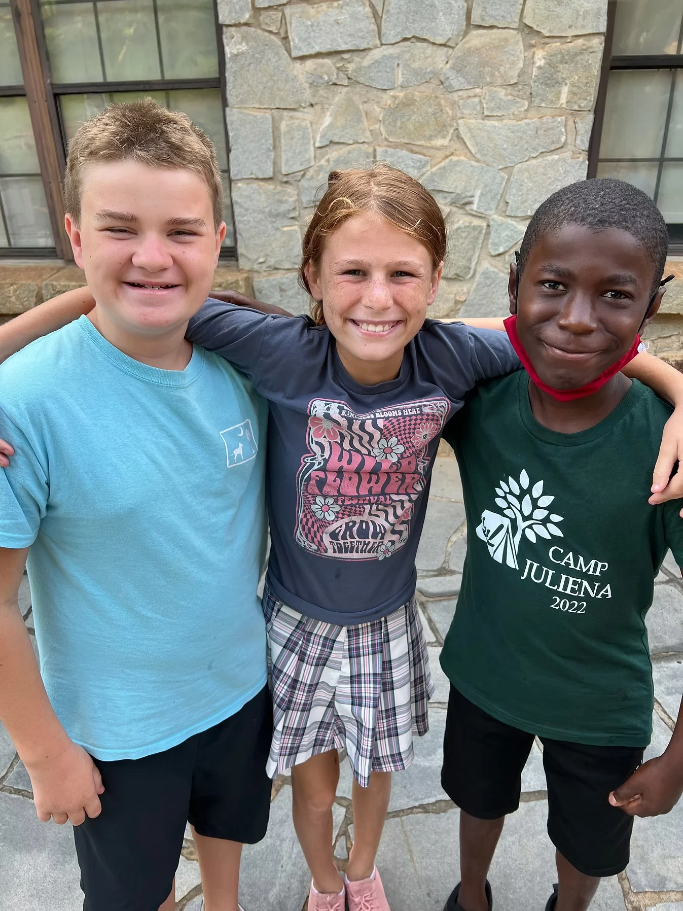 Three smiling kids, a boy and two girls, stand close together with arms around each other. One boy wears a Camp Juliena shirt.
