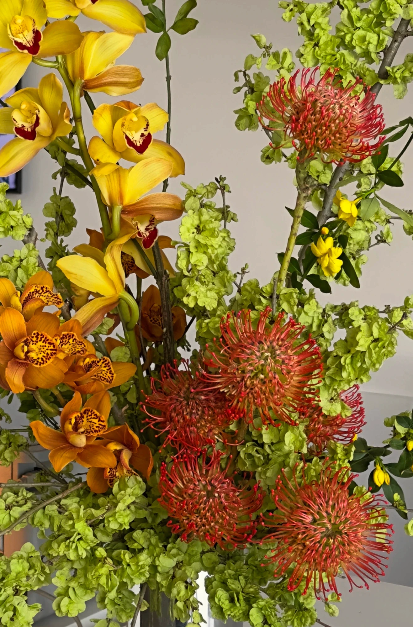Mesa decorada con un centro de mesa floral de hortensias rosadas, flores blancas y amaranto rojo colgante, acompañado de berenjenas, repollo y hojas verdes sobre un mantel blanco bordado, en una sala elegante con espejo antiguo y detalles dorados.