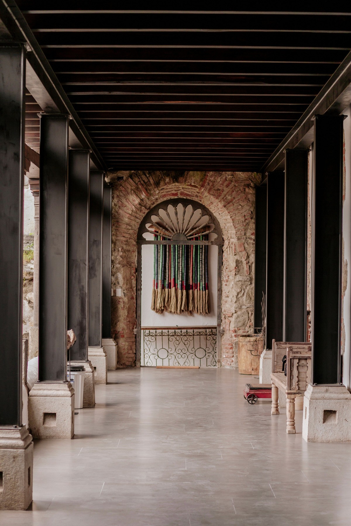 A historic architectural corridor with stone columns and a vaulted ceiling leads to an intricately carved archway adorned with a decorative tapestry.