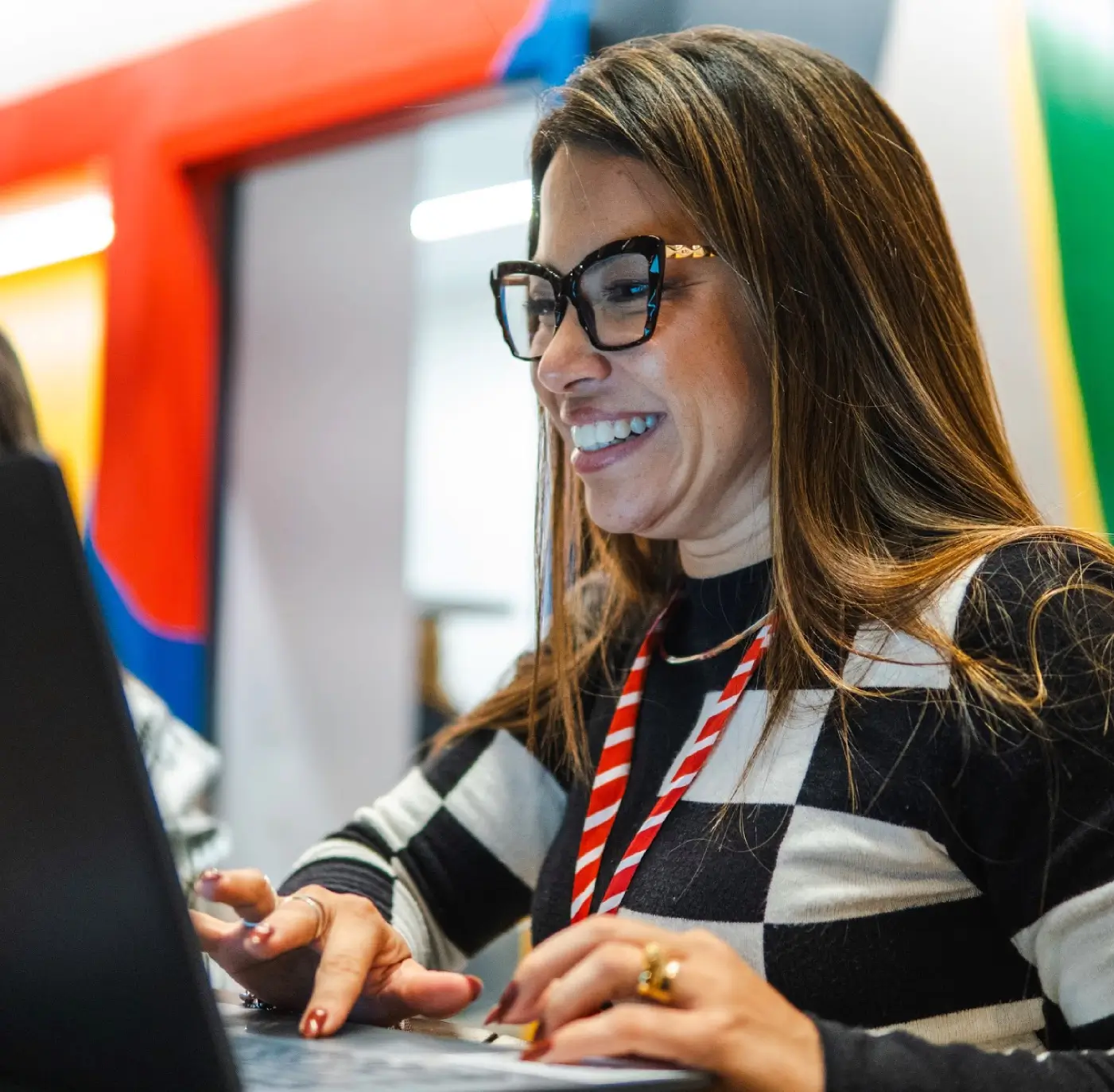 Uma pessoa usando óculos sorri enquanto digita em um laptop em um espaço de trabalho colorido.