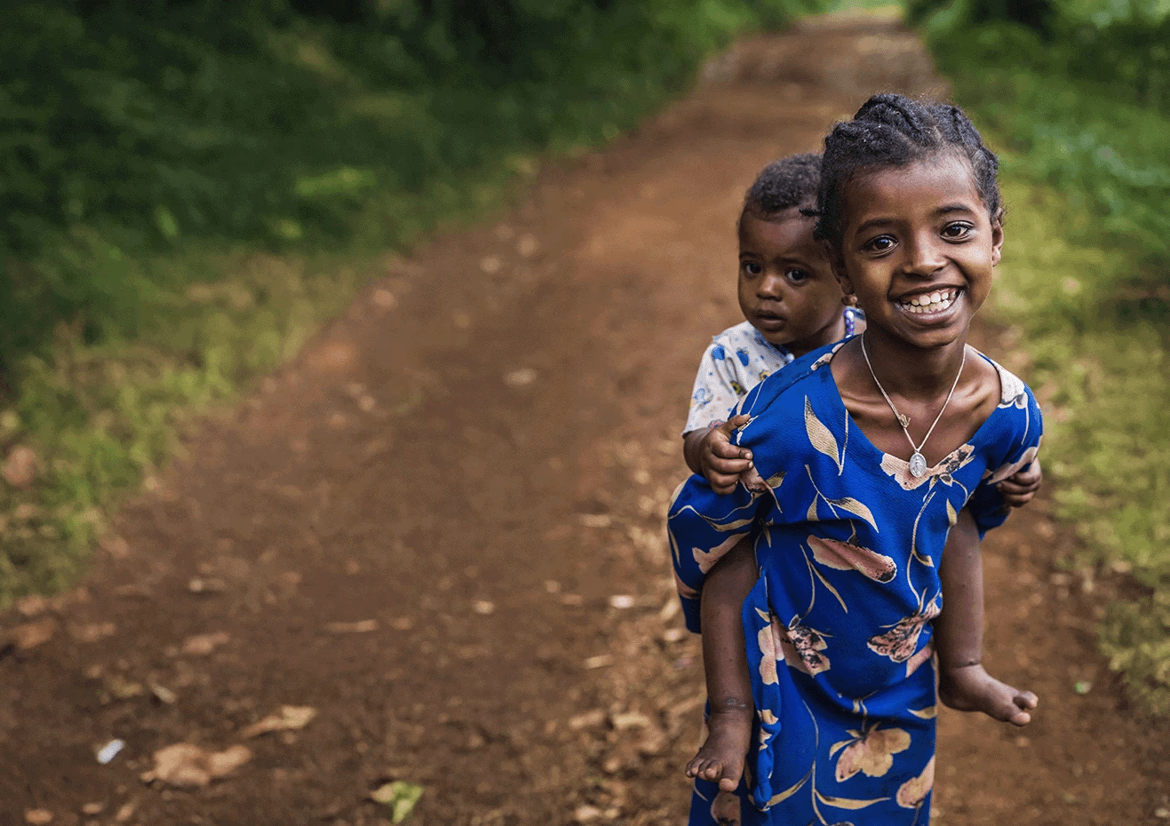 A young girl in a bright blue floral dress smiles joyfully at the camera while carrying a smaller child on her back along a dirt path surrounded by lush green foliage.