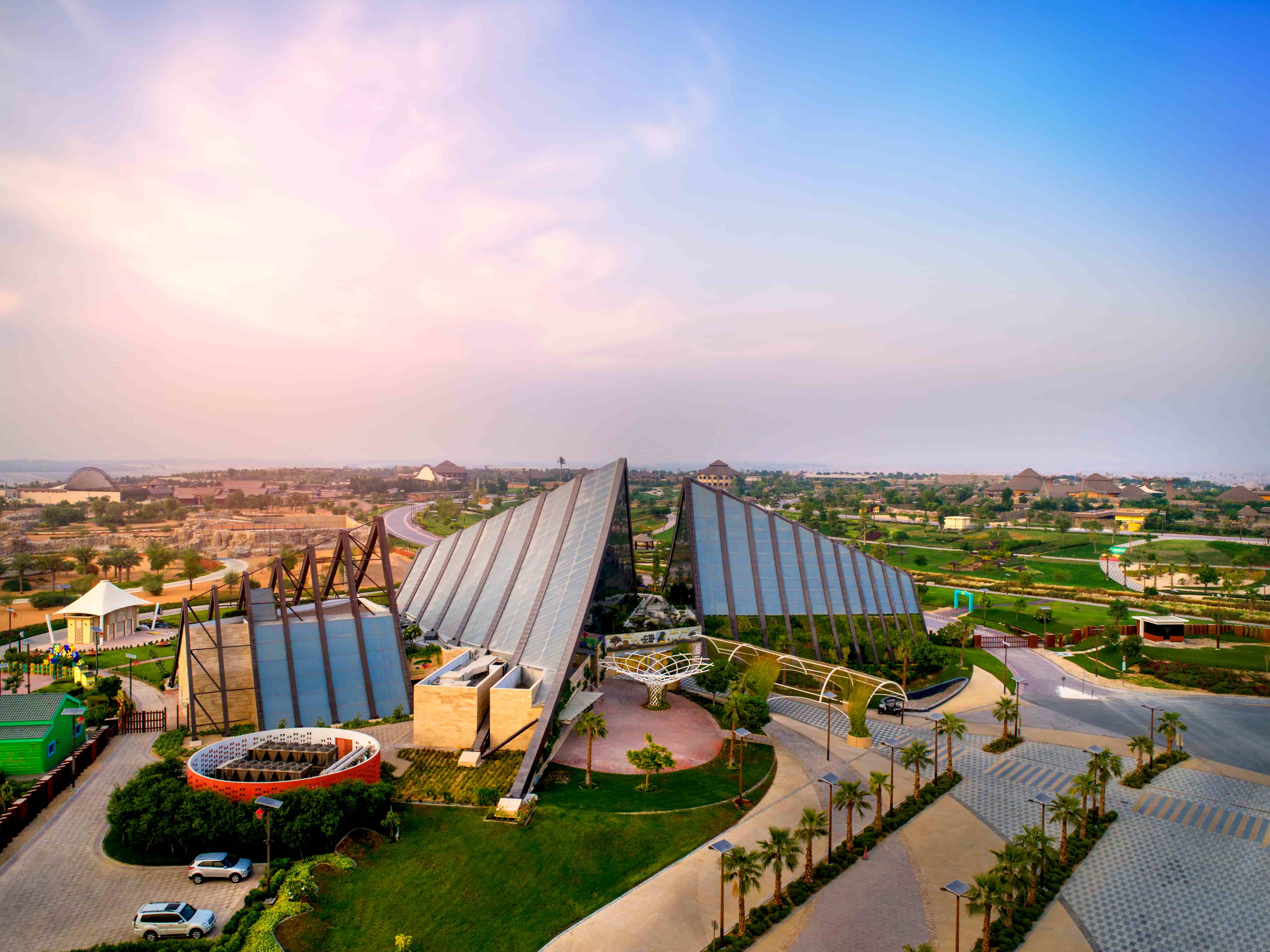 An aerial view of the Dubai Safari Park with a modern, angular glass-roof structure surrounded by greenery and pathways.&nbsp;