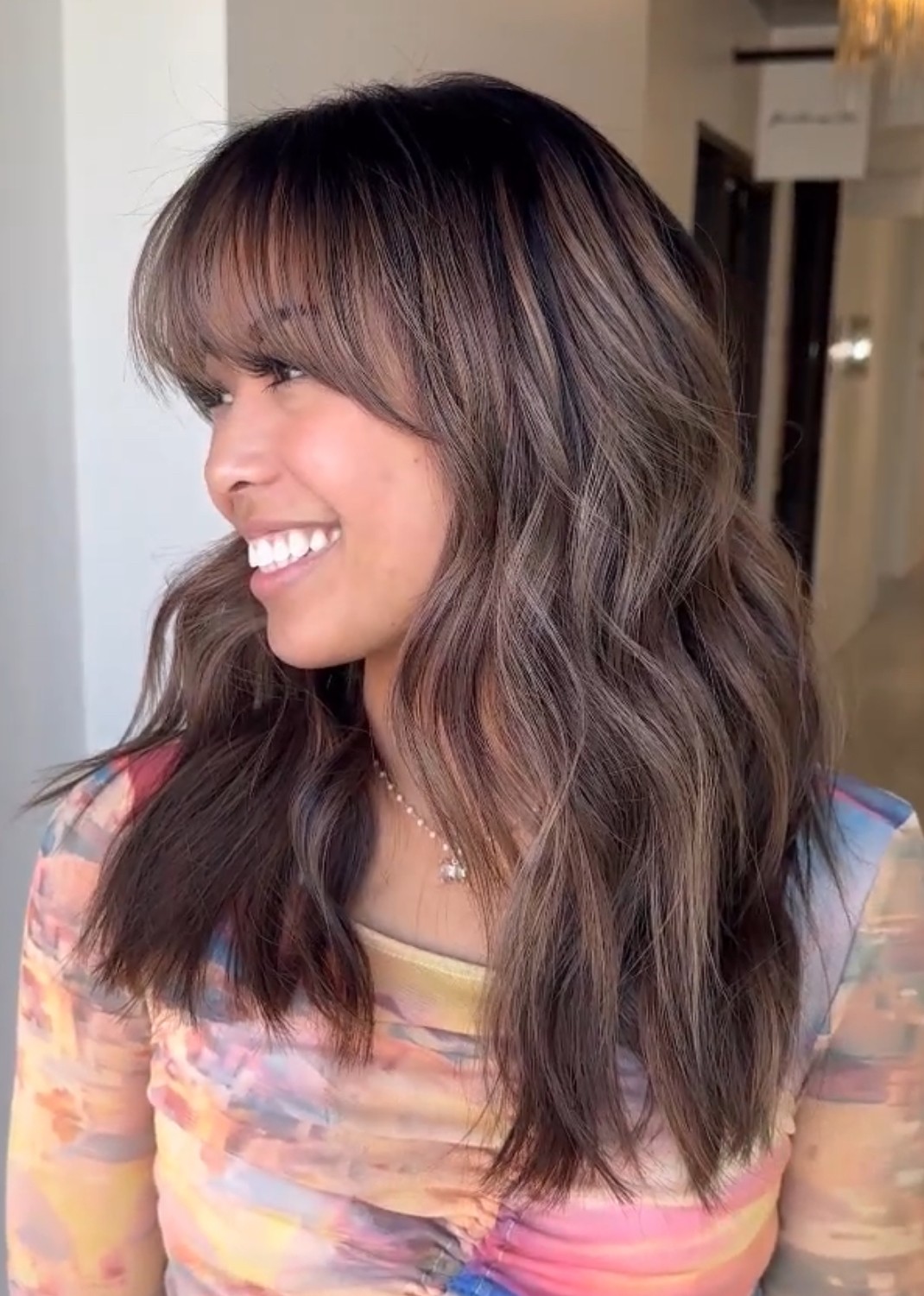 Side view of medium-length brunette hair with soft face-framing layers and wispy bangs, styled in relaxed beachy waves for an effortless finish.