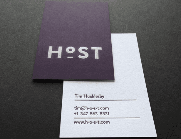 Two business cards rest on a dark gray surface. The top card is dark purple with the word "HOST" in light gray sans-serif font. The bottom card is white with black text reading "Tim Hucklesby," followed by an email address, phone number, and website address.