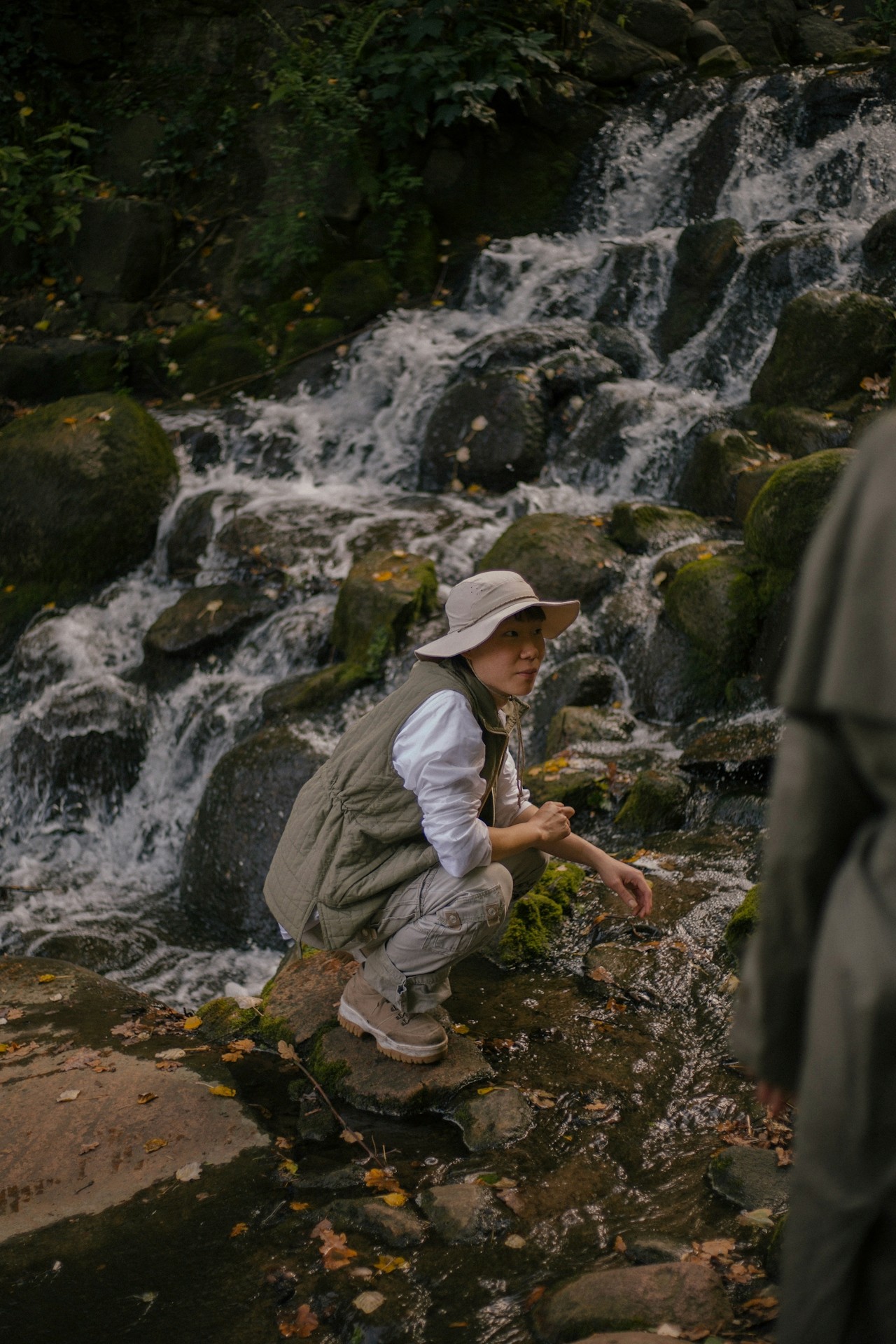 A solitary individual sitting on their knees on a rock ledge, close to where a small stream of water flows over a mountain cliff.