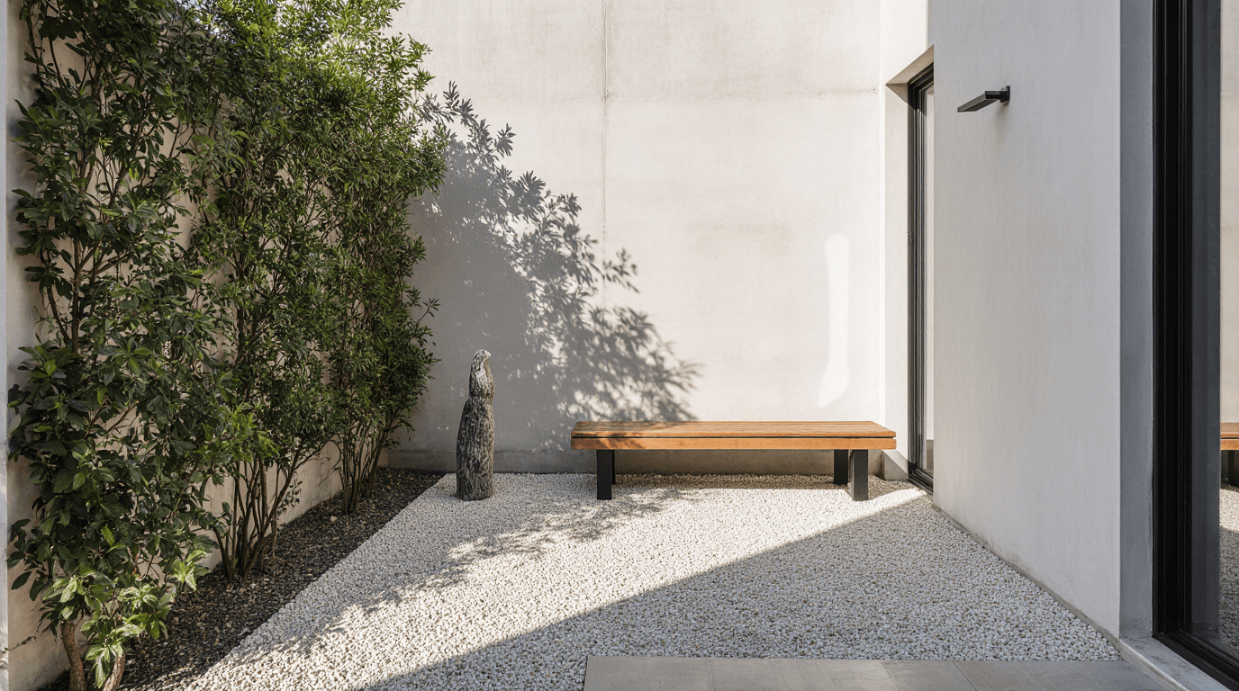 Minimalist courtyard with a wooden bench, small standing sculpture, and neatly trimmed greenery along a light-colored wall.