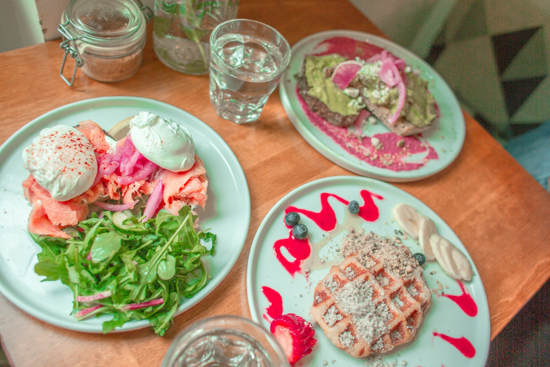 An overhead view of three breakfast or brunch plates on a wooden table. The plates feature: a savory dish with smoked salmon, poached eggs, pickled onions, and greens; a sweet plate with a waffle, bananas, blueberries, and a pink drizzle; and a third plate with avocado toast topped with goat cheese and pickled onions, drizzled with a pink sauce.