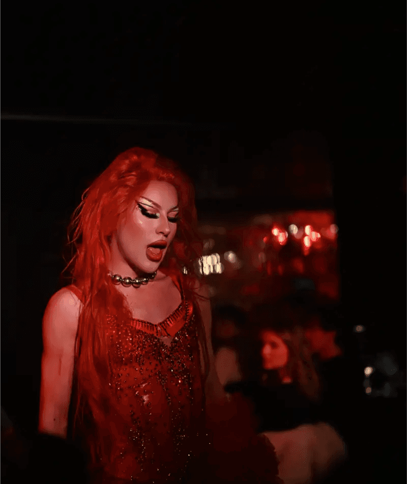 A striking close-up portrait of a glamorous drag queen with shimmering red hair and bold glitter makeup under warm cabaret spotlights.