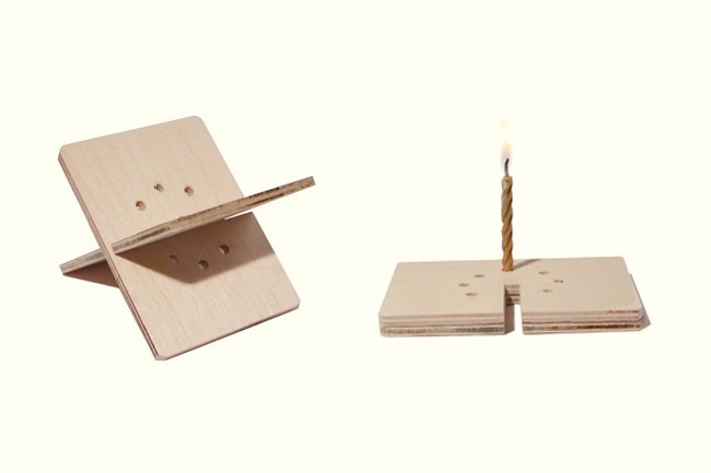 A light-colored wooden candle holder is positioned on the left side of the image. It has a square base with four small holes and a horizontal piece extending outward. A single lit candle with a twisted wick is placed on a stack of similar wooden pieces to the right. The background is plain white.