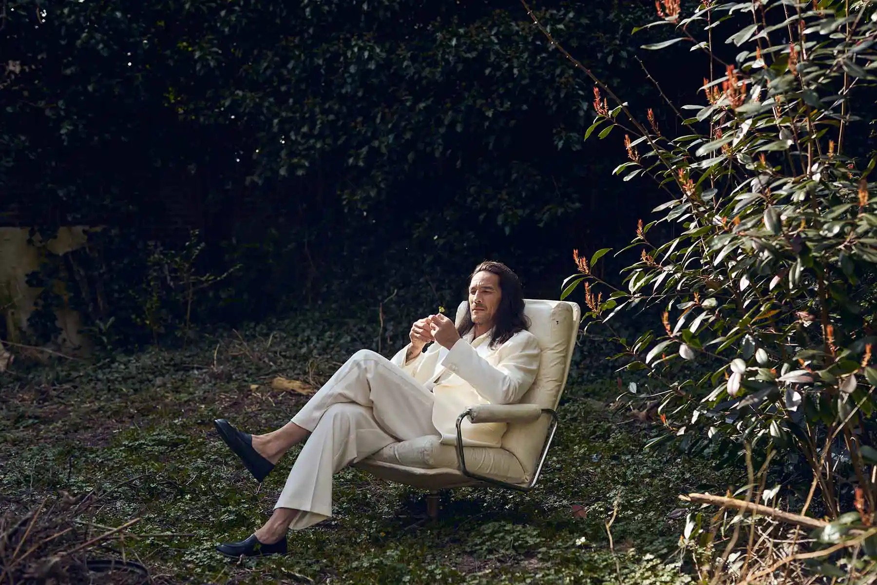 Model reclines outdoors on a garden bench in pale clothing.