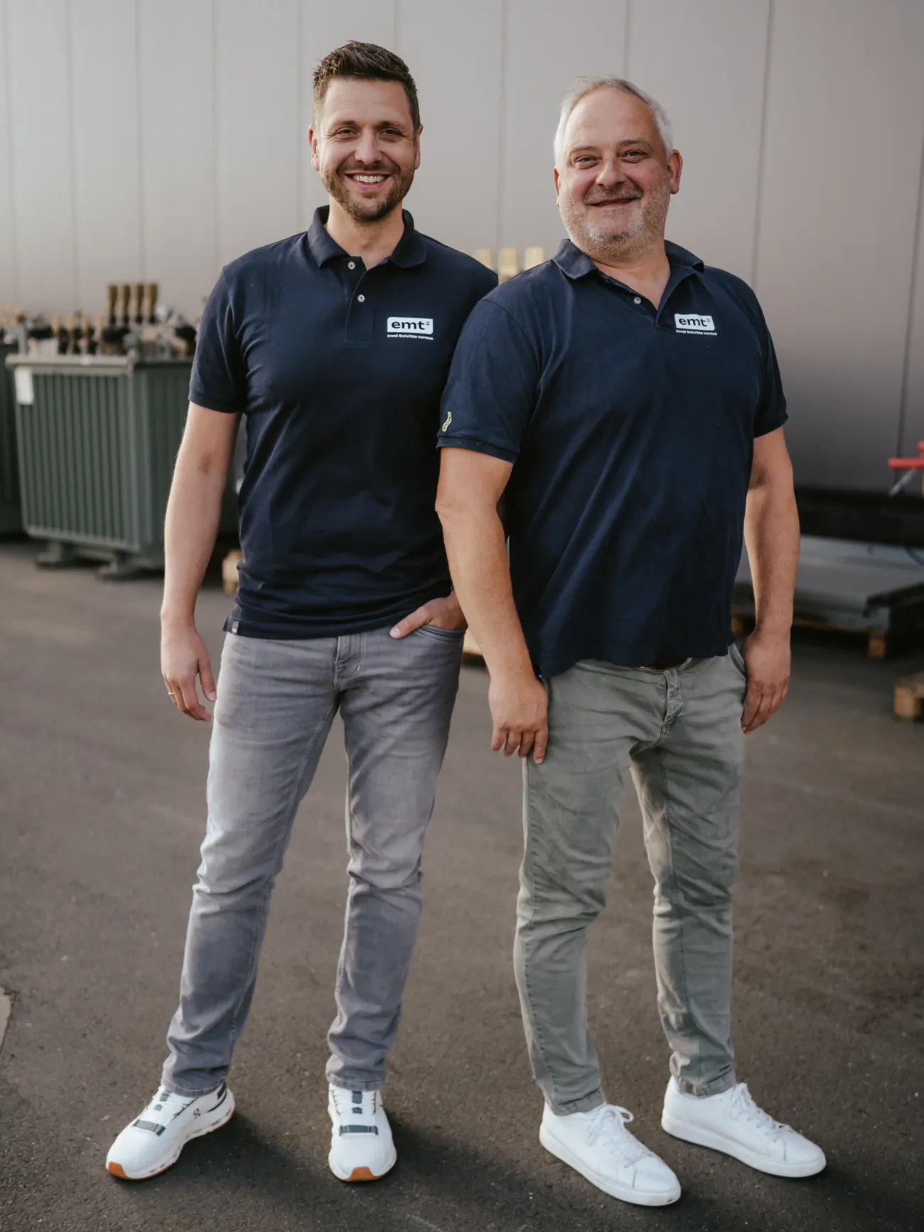 Zwei Männer in Poloshirts und Hosen vor einem Gebäude mit dem Logo „emt²“.