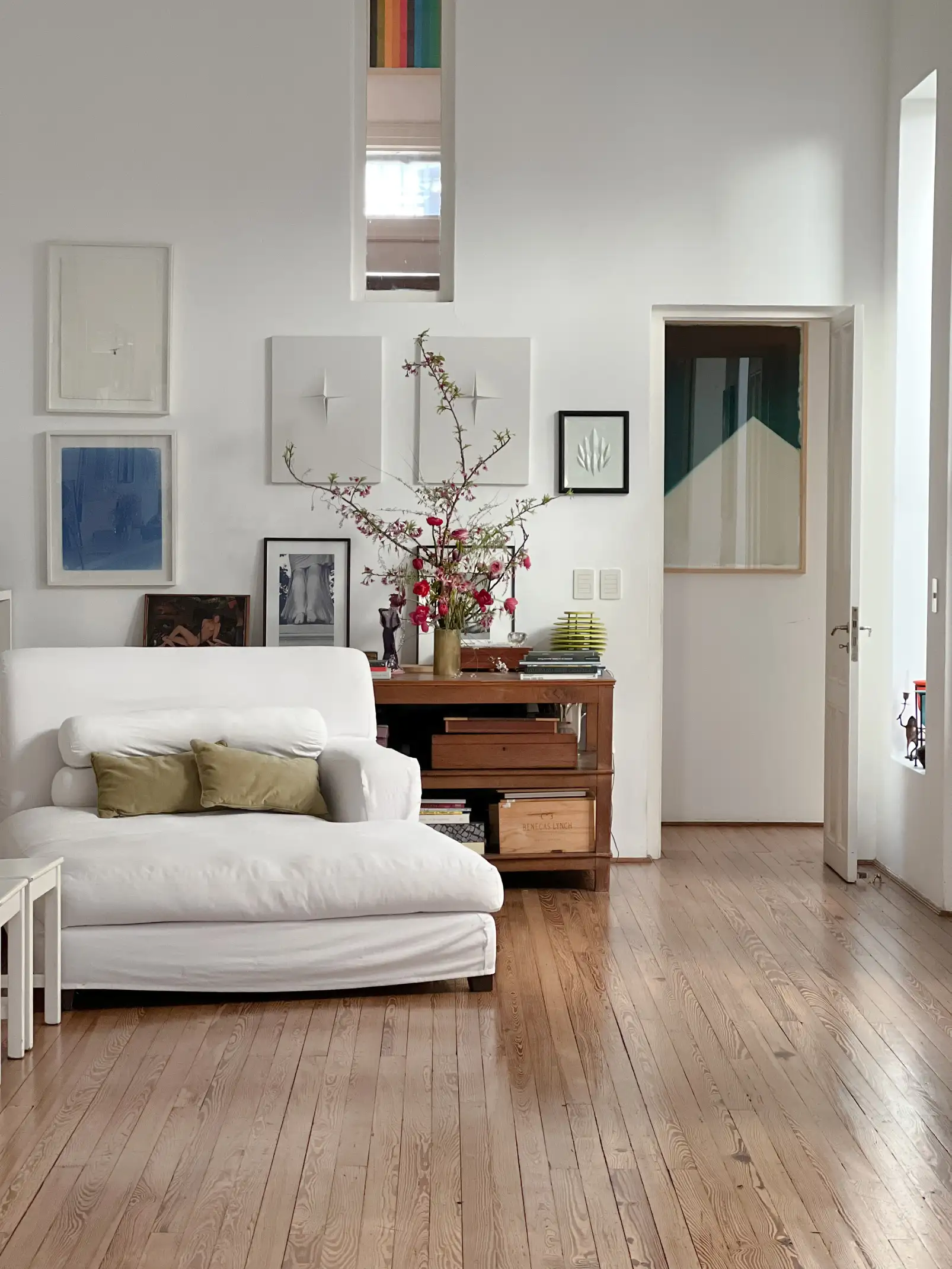 Arreglo floral con tulipanes, narcisos y lirios en tonos blanco, amarillo y rosa en un jarrón de cerámica, colocado sobre una mesa de madera junto a una lámpara cromada con pantalla blanca.