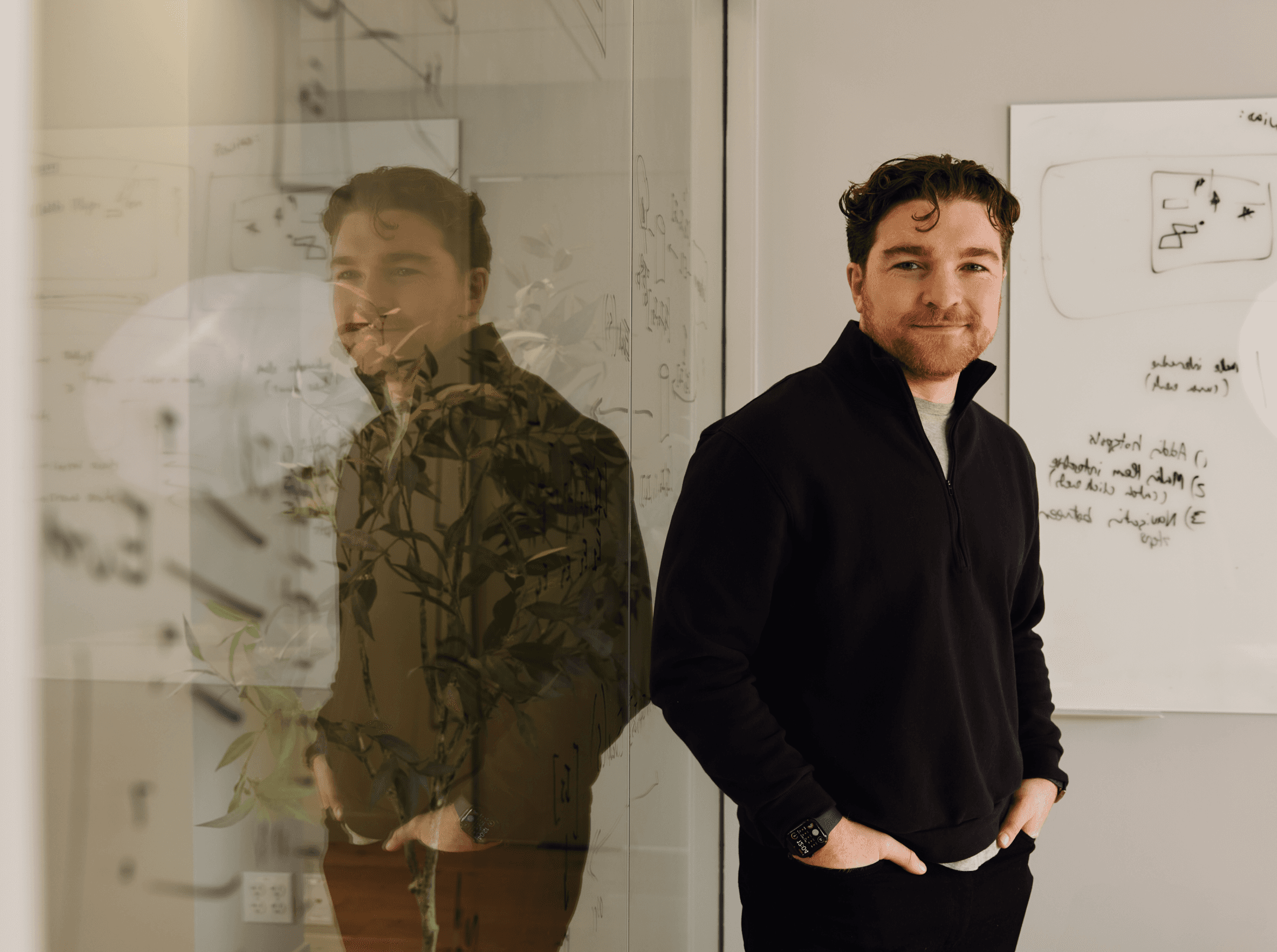 Portrait of a team member standing in a modern office beside a glass wall with whiteboard sketches and reflections in the background.
