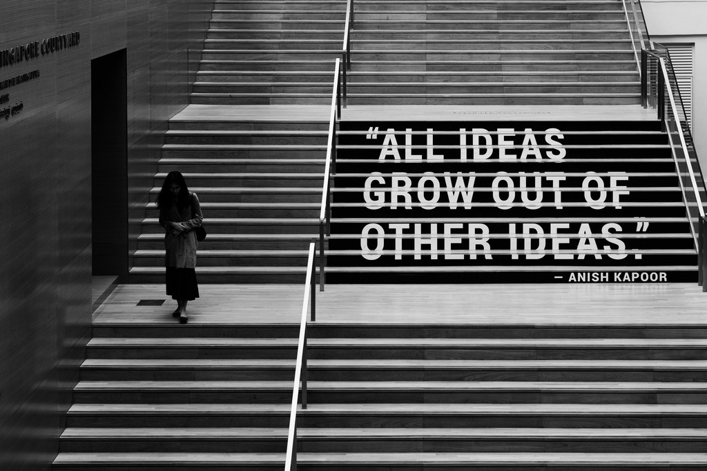 Bkacj white image taken from a ledge showing a staircase. A woman walking to the left and on the right side of the stairs is written "All ideas grow out of other ideas"."