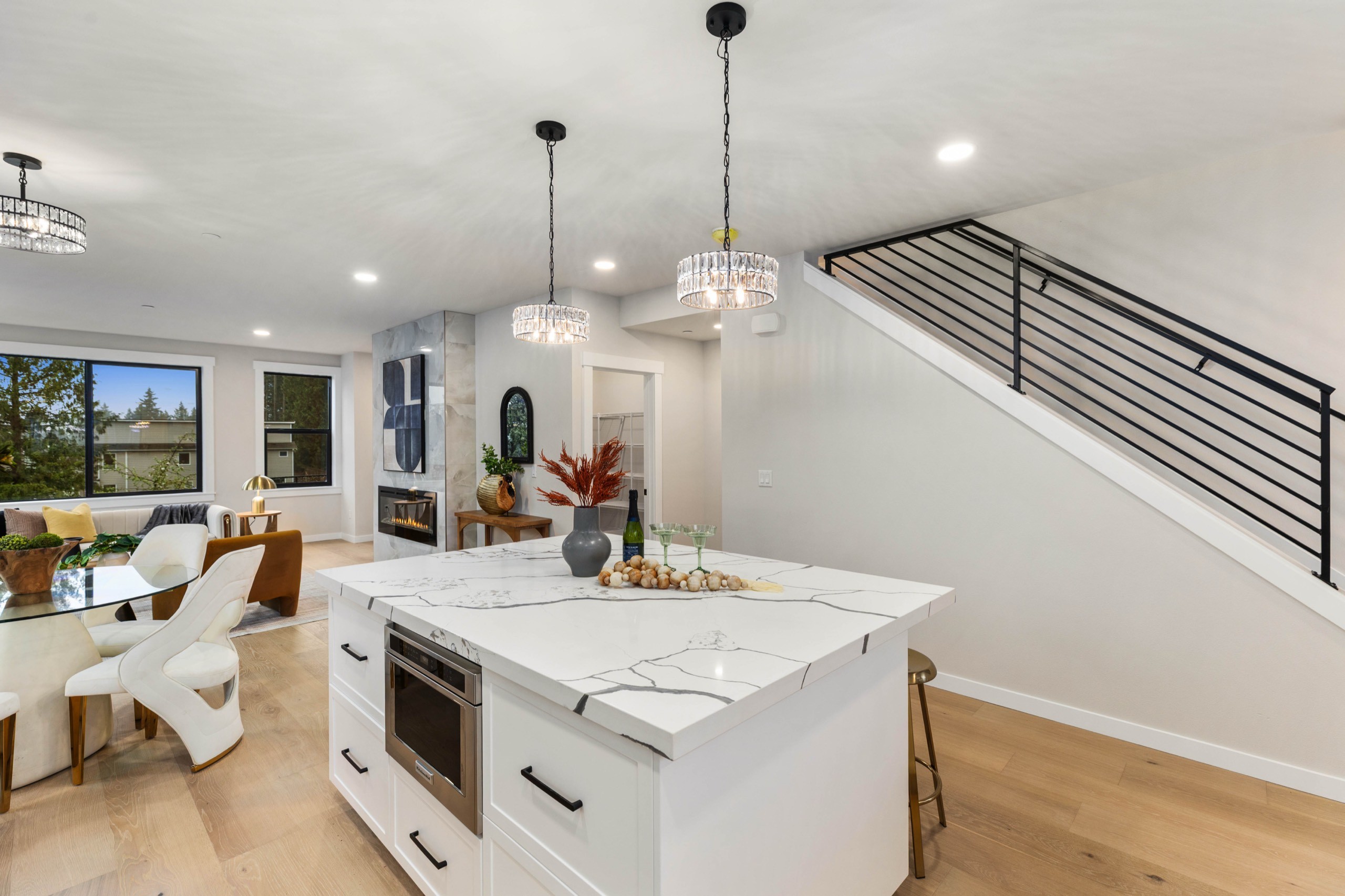 Luxury open-plan kitchen with large white waterfall island, pendant lighting, microwave drawer, marble countertops, and black metal stair railing in background