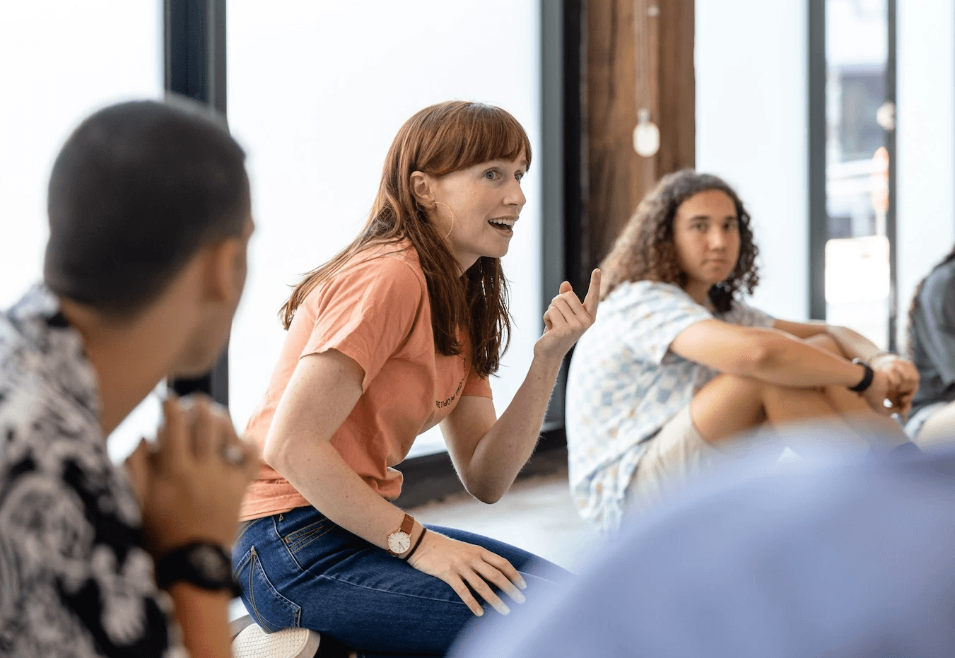A facilitator kneels on the floor speaking animatedly to a group of young people seated in a circle during a workshop, in a bright studio space with large windows.