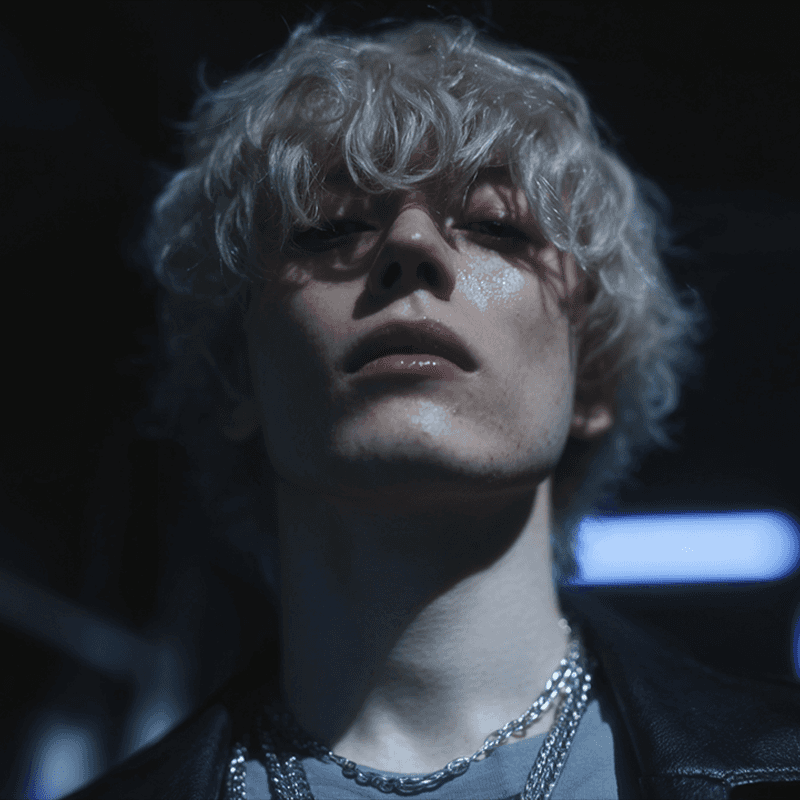 Young person with tousled blonde hair and a thoughtful expression, lit dramatically from above. Wears a dark jacket and chain necklace, suggesting a moody, introspective vibe.