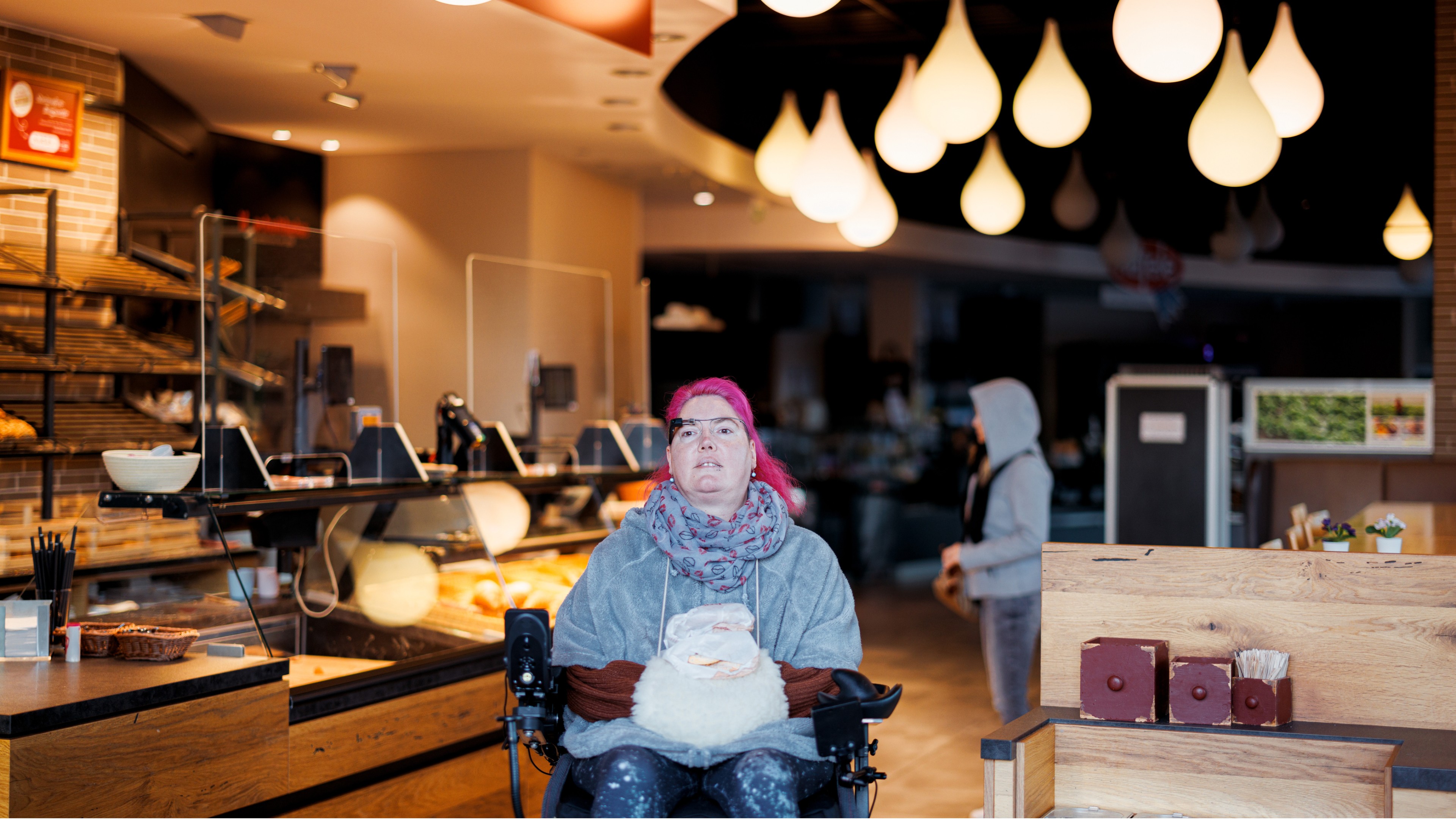 Saskia, a user of the munevo smart glasses, sitting in a motorized wheelchair inside a café. She has striking pink hair and is wearing a grey scarf and a light grey jacket. Saskia is looking forward, using the smart glasses to navigate or interact with her environment. The café setting is warmly lit, with stylish hanging lights in the background, creating a cozy and inviting atmosphere. This setting emphasizes the everyday usability of the munevo glasses, enabling mobility and independence for individuals in various social settings