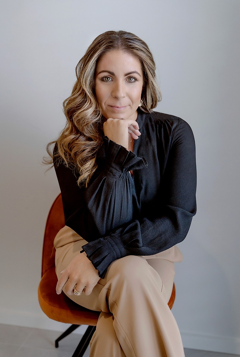 Minimal studio portrait of a confident woman in a white shirt, arms crossed, on a soft gray background.