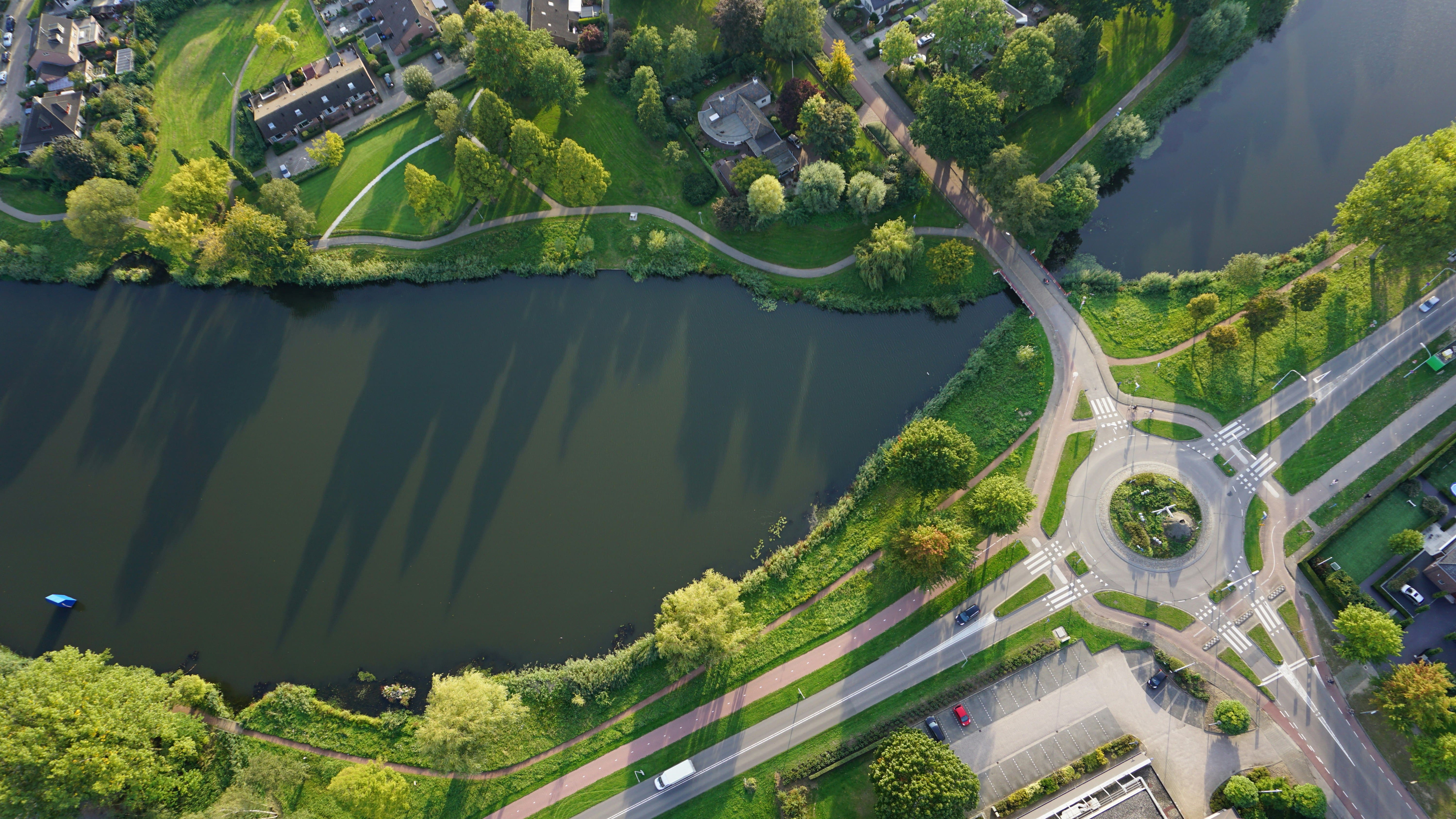 Luchtfoto van een woonwijk met donker water, groen gras, bomen, huizen en een rotonde met wegen en fietspaden.