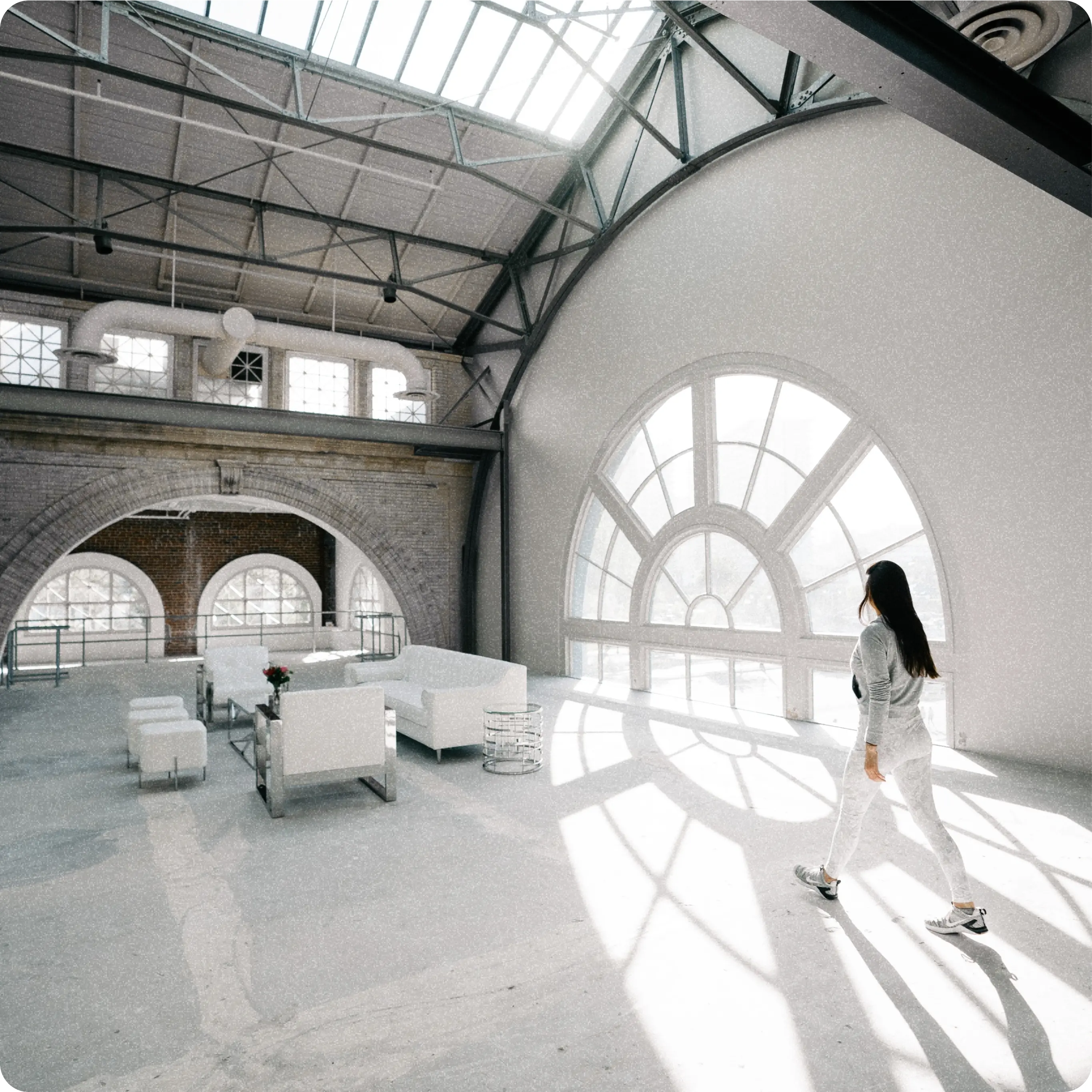 An interior view of a spacious, bright loft studio with large arched windows and a high, industrial-style ceiling with metal beams. The room is furnished with modern white sofas and chairs. A woman dressed in white is walking towards the large window, which allows abundant natural light to flood the space, creating a serene and open atmosphere.