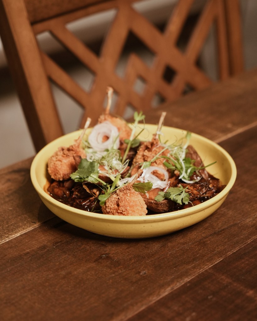 A yellow bowl filled with a contemporary take on three cup frog dish, featuring crispy items garnished with green herbs and sliced onions, placed on a wooden table.