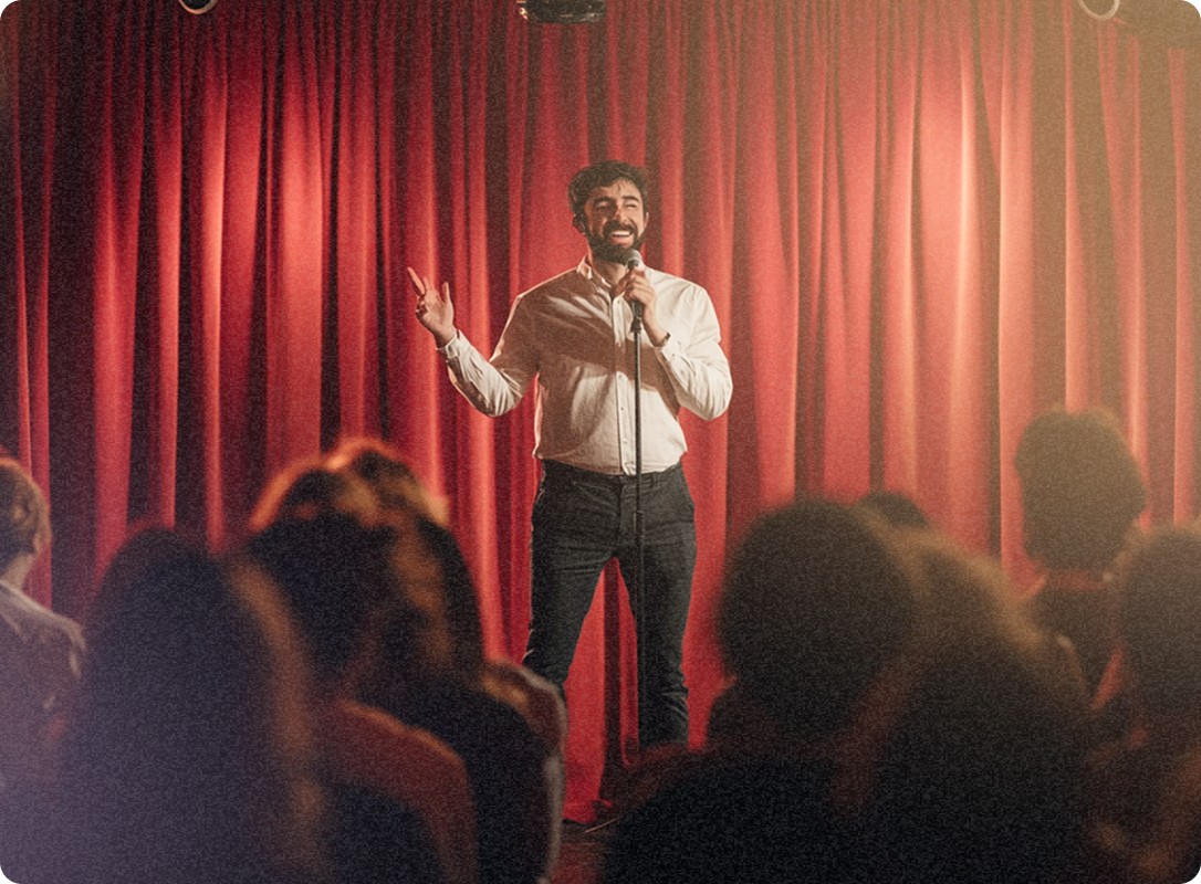 Un comédien sur scène, vêtu d'une chemise blanche, se tient devant un rideau rouge en train de faire une performance, avec un public qui l'écoute attentivement.