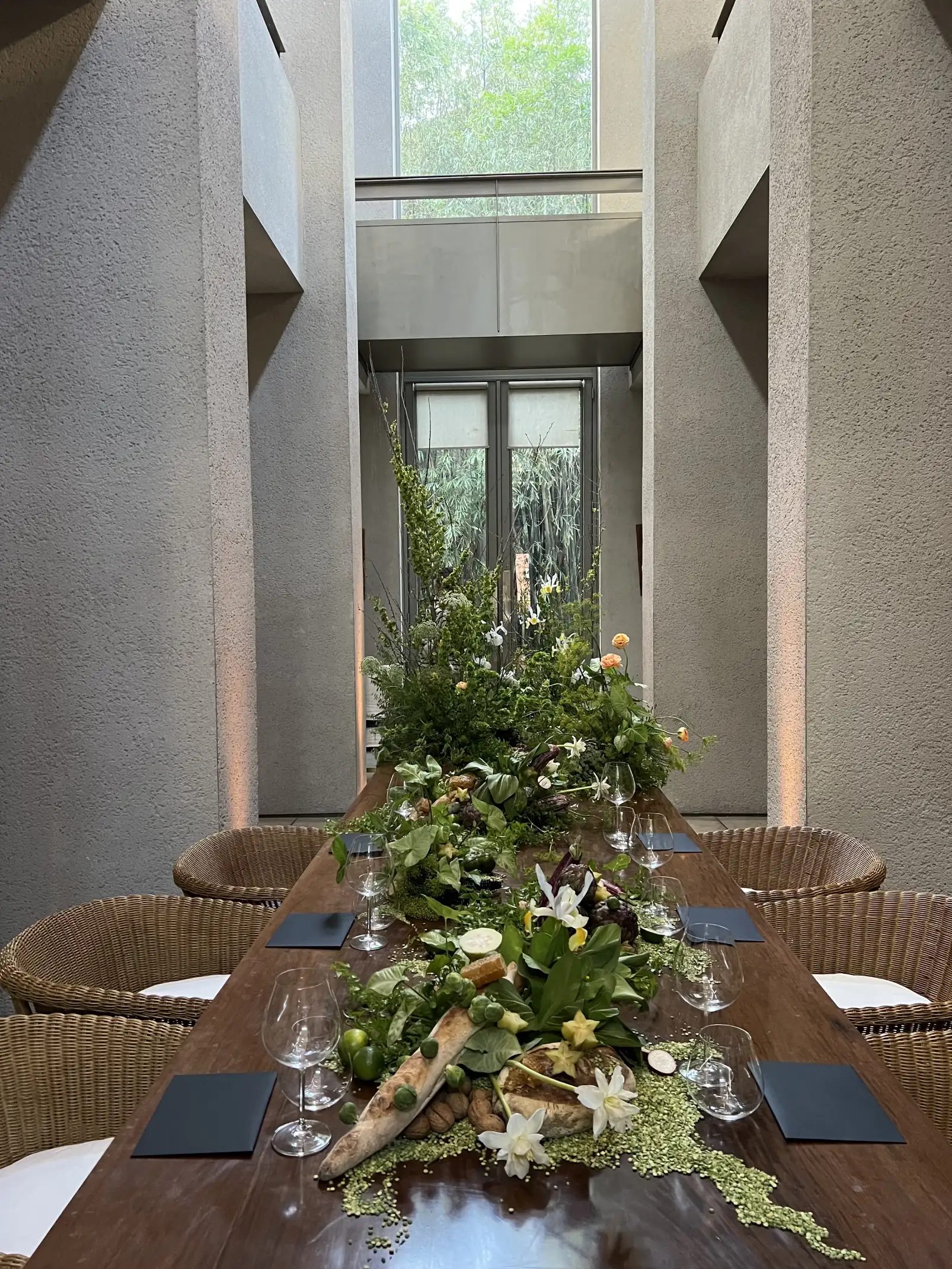 Vista frontal de mesa larga decorada con elementos naturales, flores y alimentos en entorno arquitectónico minimalista.
