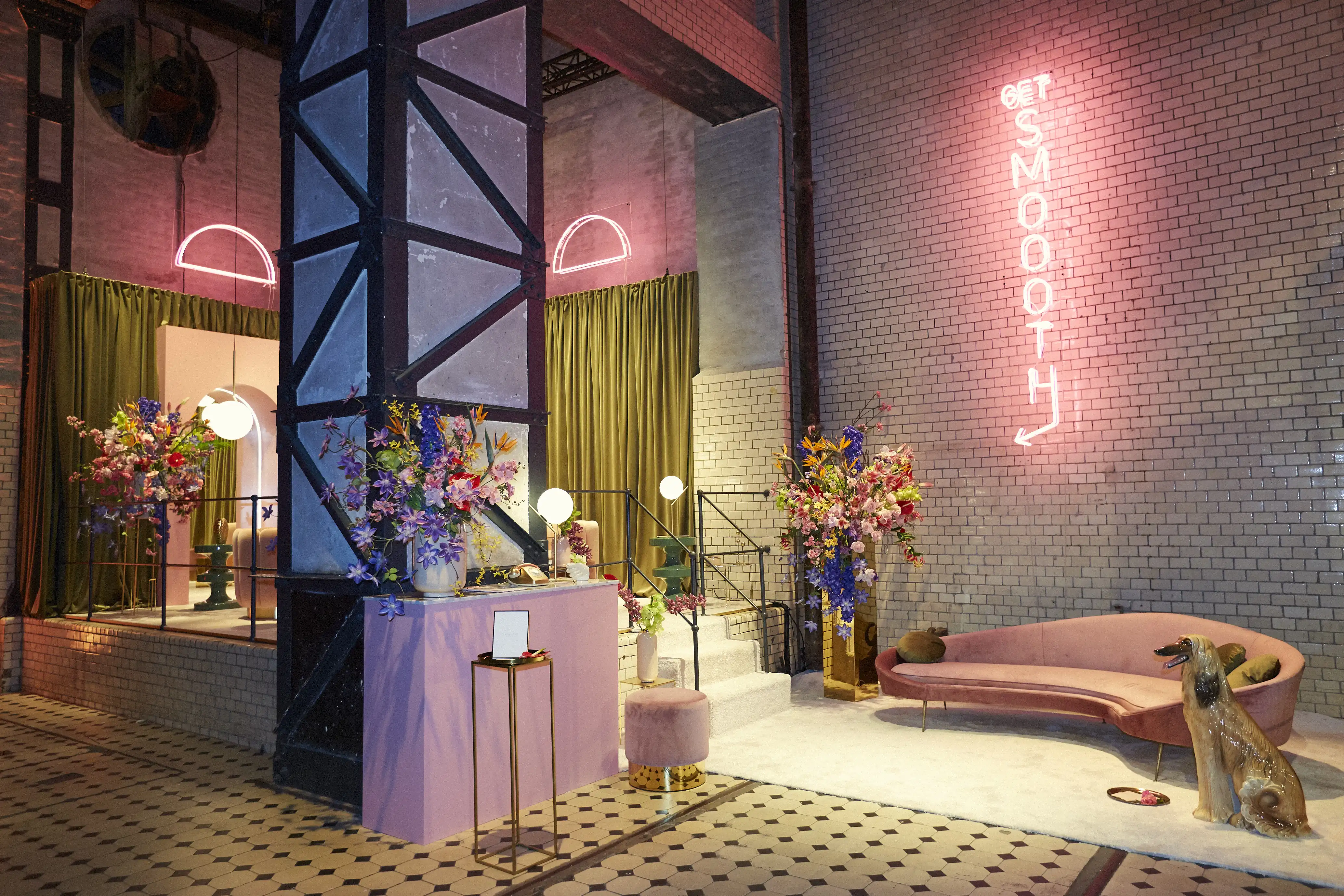 Industrial hall with steel columns transformed into a pink Klarna lounge with neon arches, flower towers, velvet sofa and a dog.