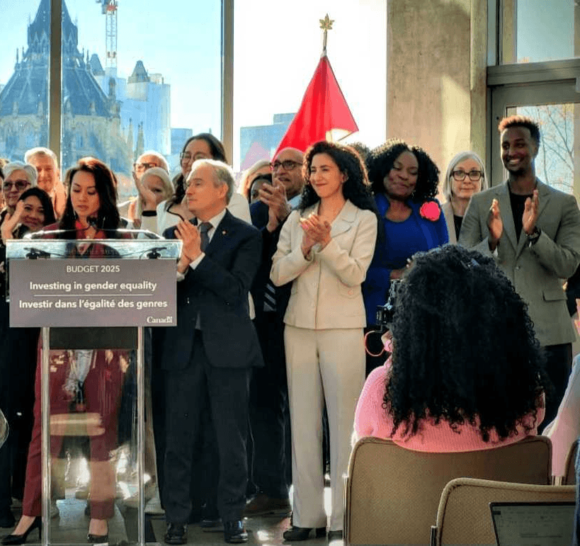 A group of people standing at a podium, smiling and applauding, with a large window in the background.