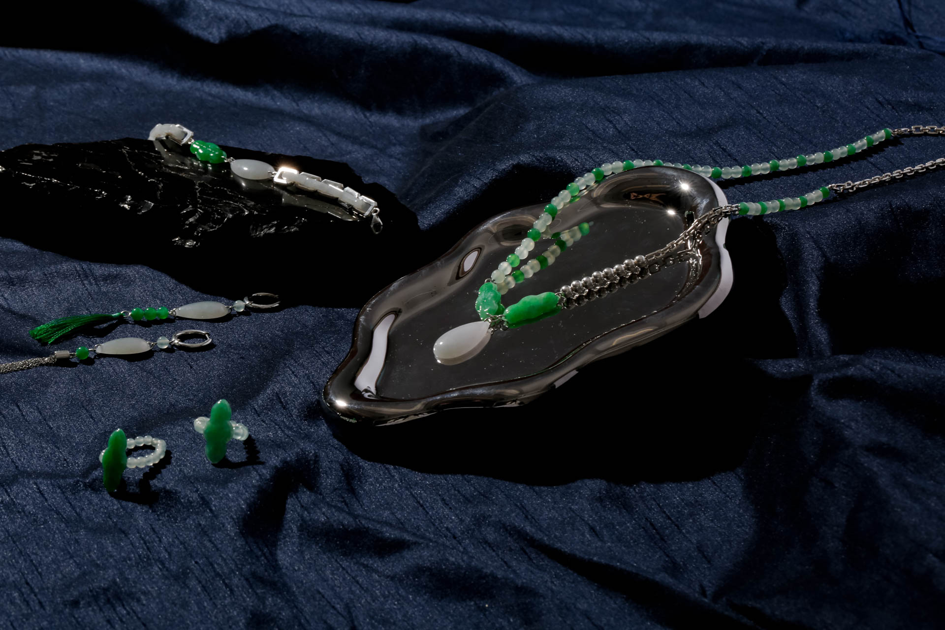 A necklace with green and white beads rests on a dark, textured surface. The beads are strung together on thin green cords that extend beyond the frame. A silver pendant, shaped like a dish, lies on the dark surface near the necklace. The pendant reflects light, and its surface appears smooth and polished. The dark background contrasts with the necklace and pendant, making them stand out prominently.