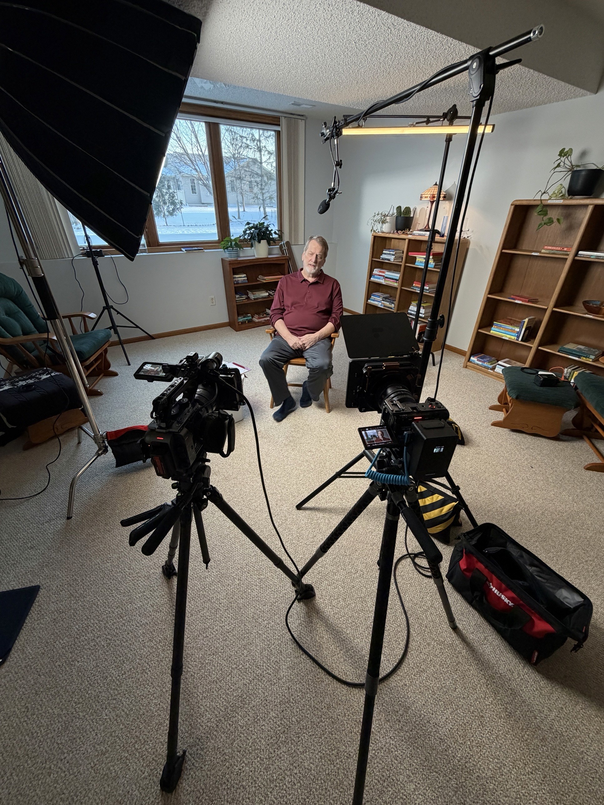 Two-camera interview setup in a basement den, the subject seated by a window with snow outside