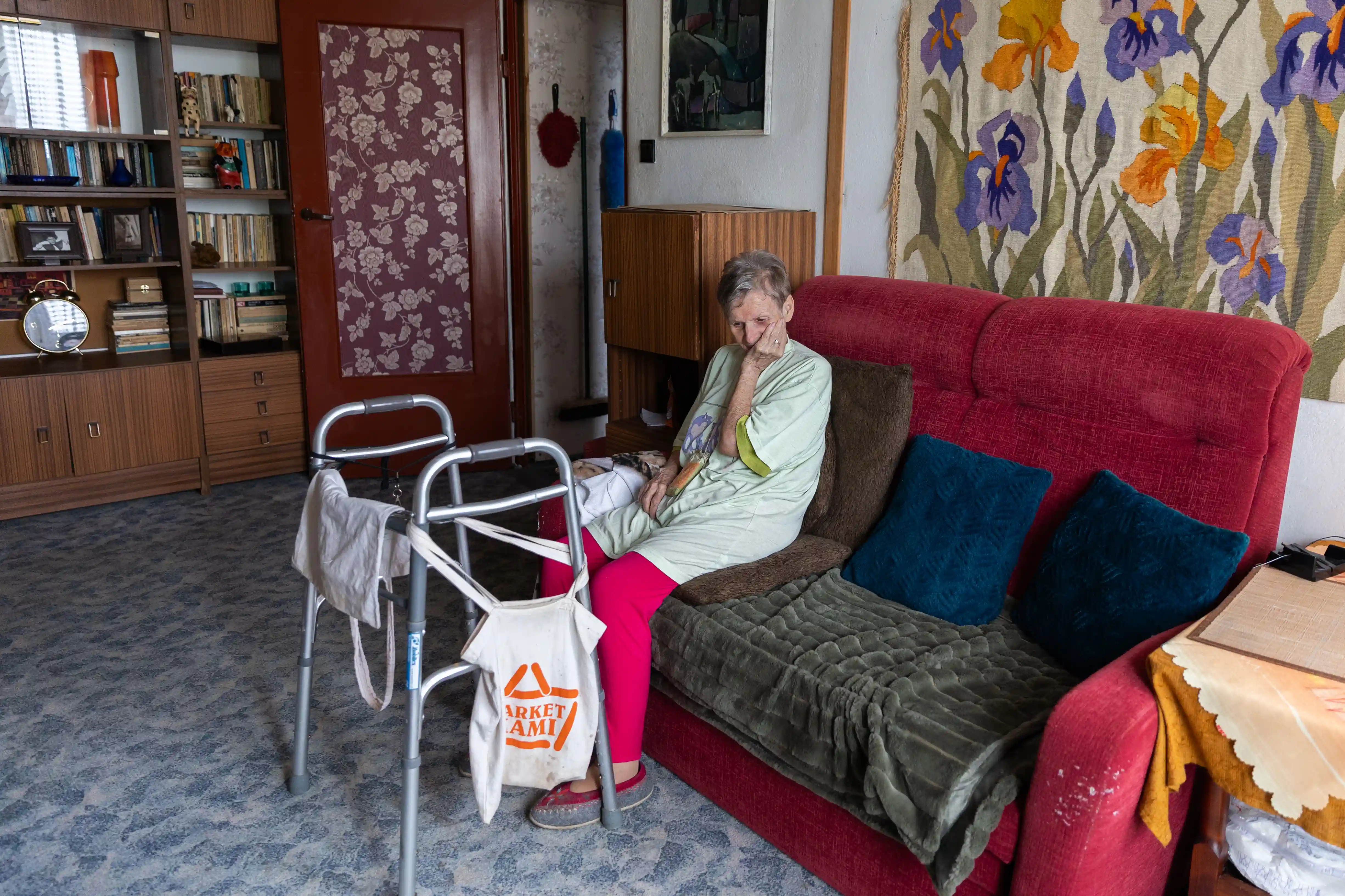 Starsza kobieta siedząca na fotelu, podpierająca głowę ze zmartwieniem. Przed nią stoi chodzik z zawieszonymi na nim siatkami.