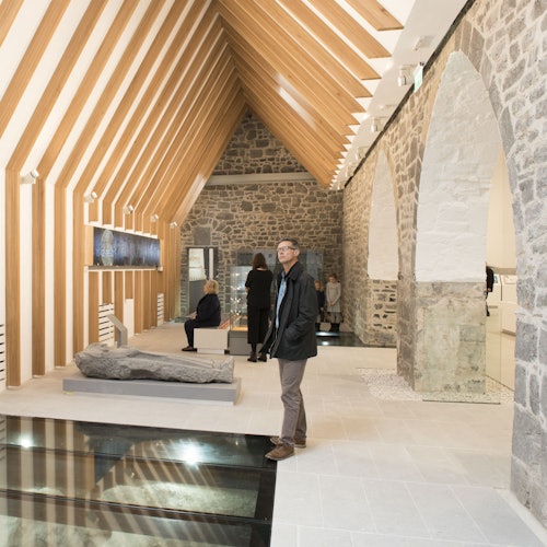 游客在一个现代化的画廊中参观，画廊有石墙、木梁、透明地板区，以及展示中的岩石雕塑。