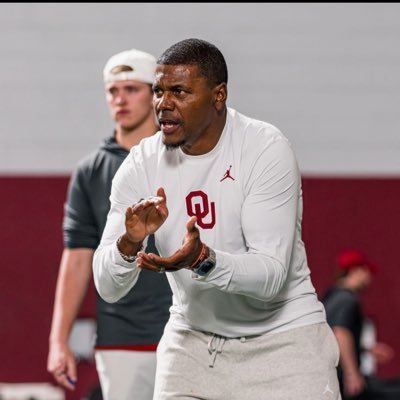 Deland McCullough in an OU shirt claps and gives instructions during practice indoors, while another person in a white cap stands in the background.