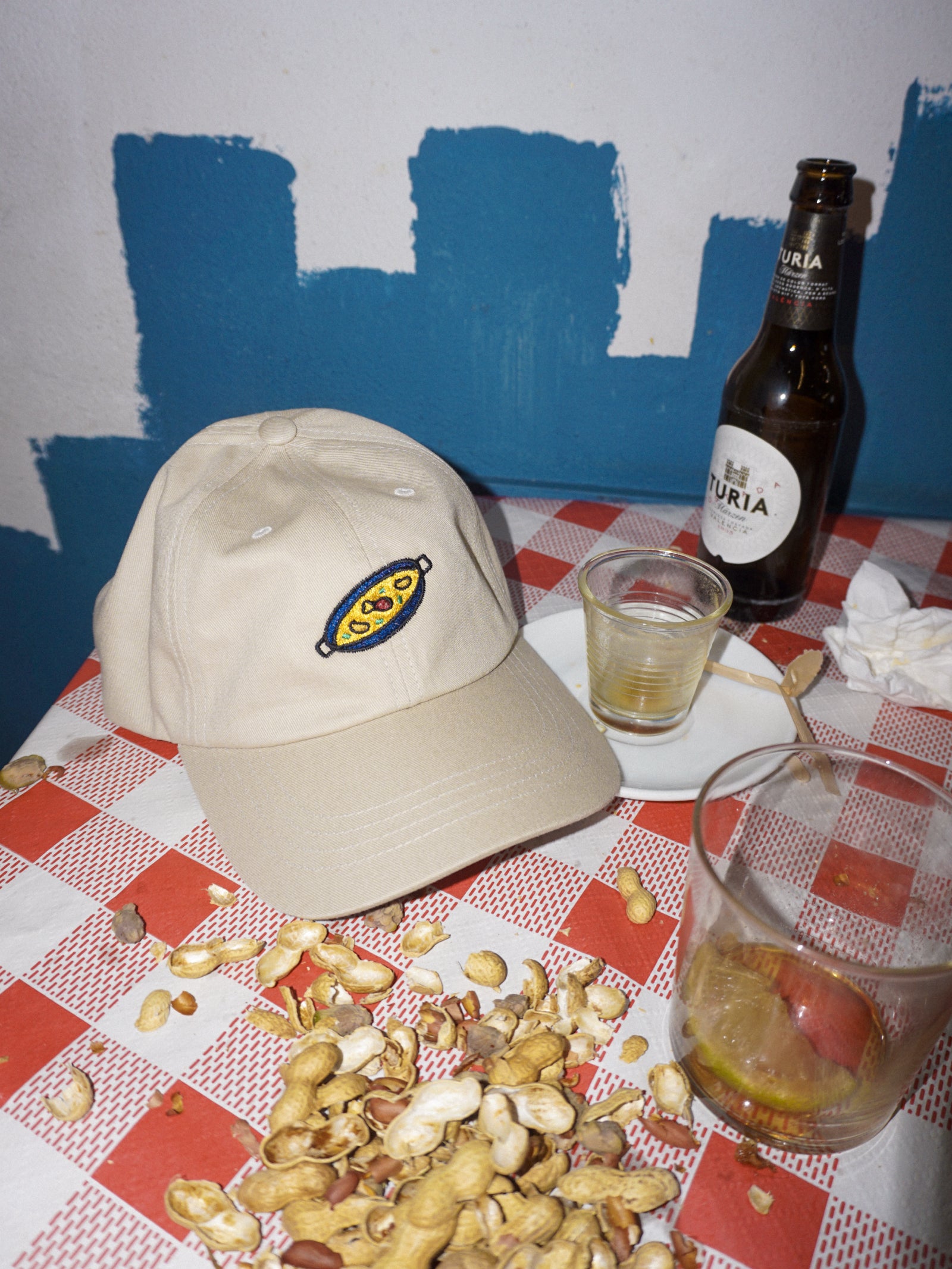Beige cap, bottle, glass, and peanuts on a checkered tablecloth with blue wall background