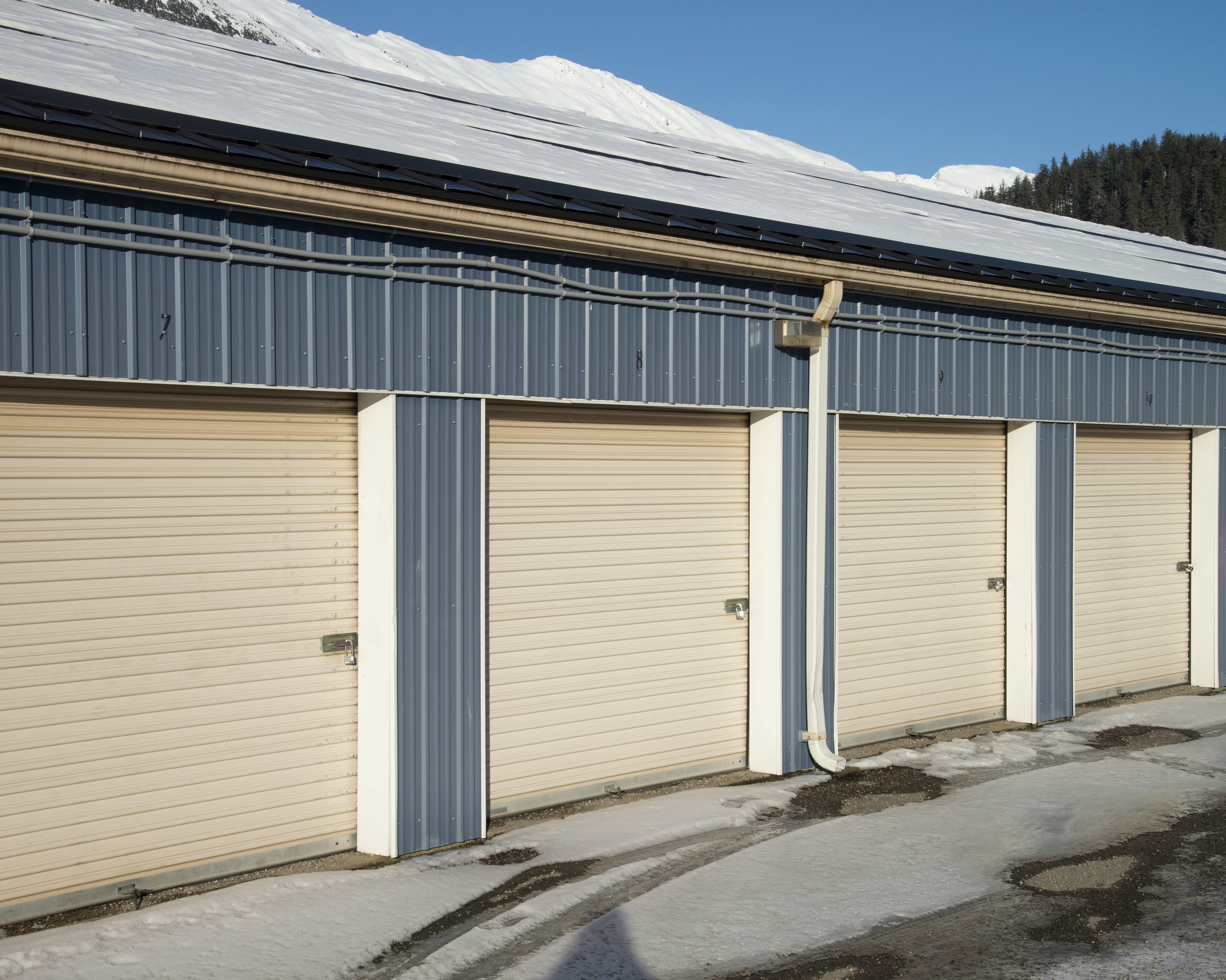 Close-up exterior view of three beige roll-up storage unit doors on a metal panel building with snow accumulated on the roofline, a downspout between units, and a clear blue sky above a snow-dusted treeline.