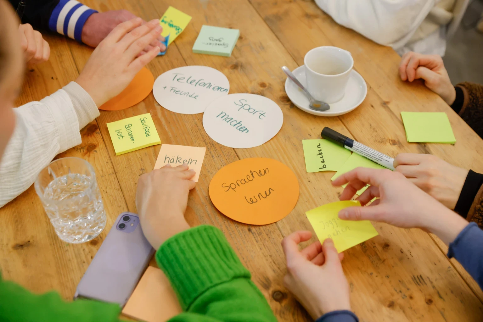 Viele Zettel und Post Its liegen auf einem Tisch, es sind einige Hände von oben zu sehen, die die Zettel anfassen und sortieren