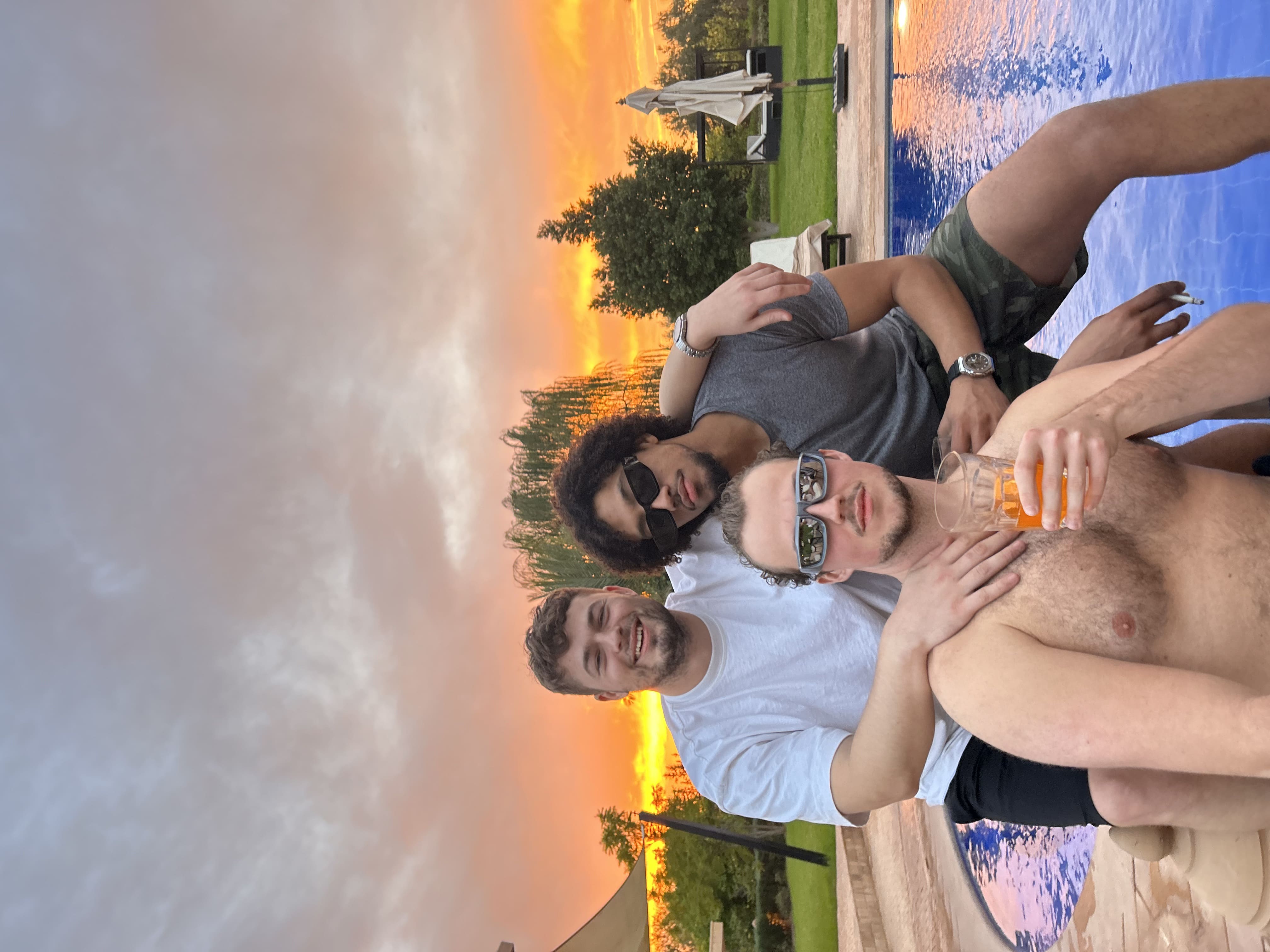 Three people smiling and enjoying drinks outdoors during sunset, with a colorful sky in the background.