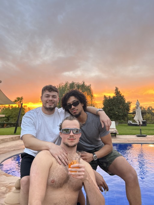 Three people smiling and enjoying drinks outdoors during sunset, with a colorful sky in the background.