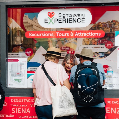 Three people reading a travel agency window display for sightseeing tours and attractions, with various brochures and signs visible.
