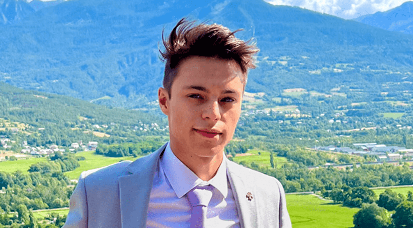 Photo de Léni Touzelet dans un costume gris, appuyé contre un petit mur en pierre