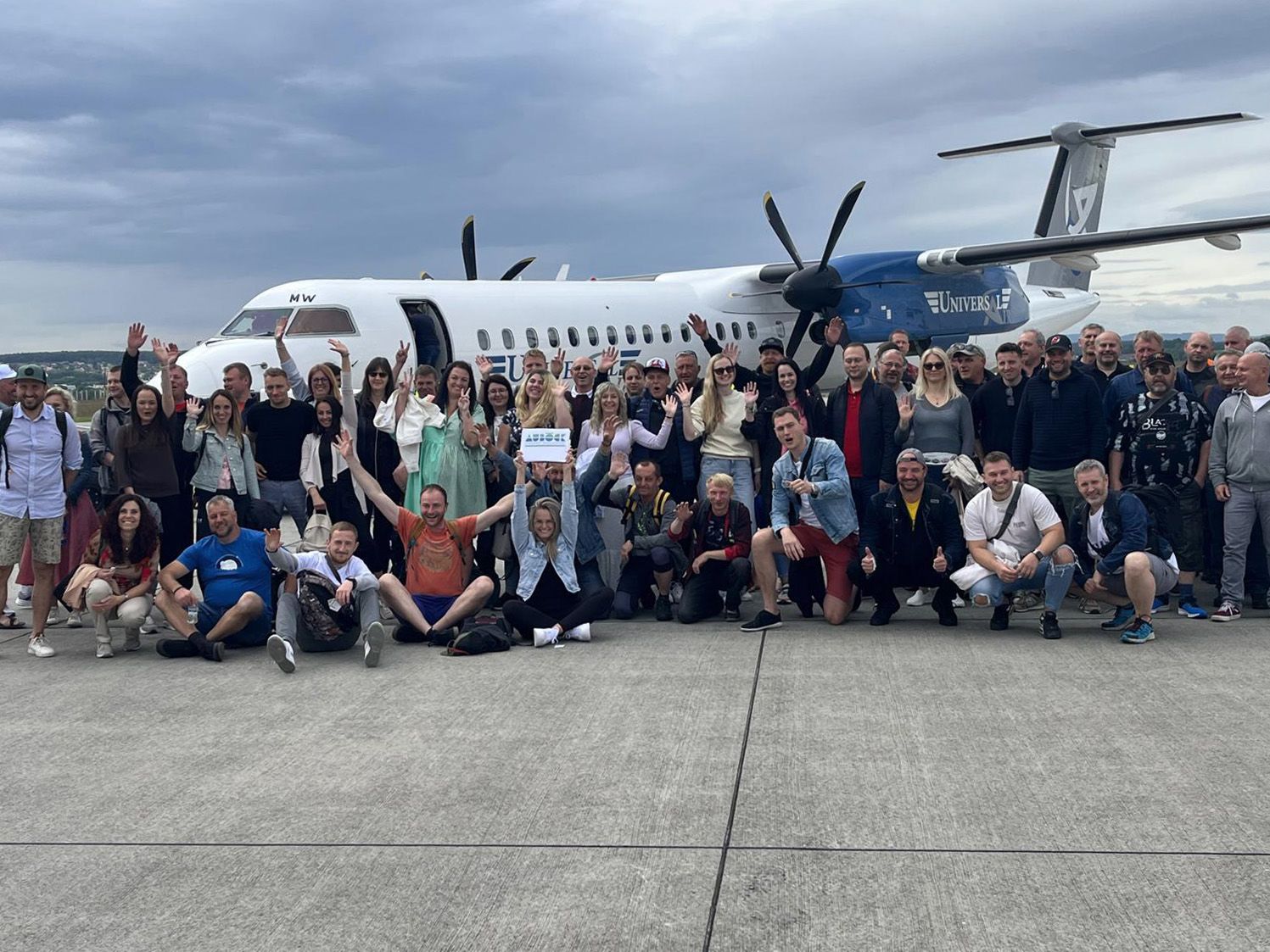 Gruppe von Personen posiert vor einem Flugzeug. Viele Personen in verschiedenen Outfits, heben die Hände.