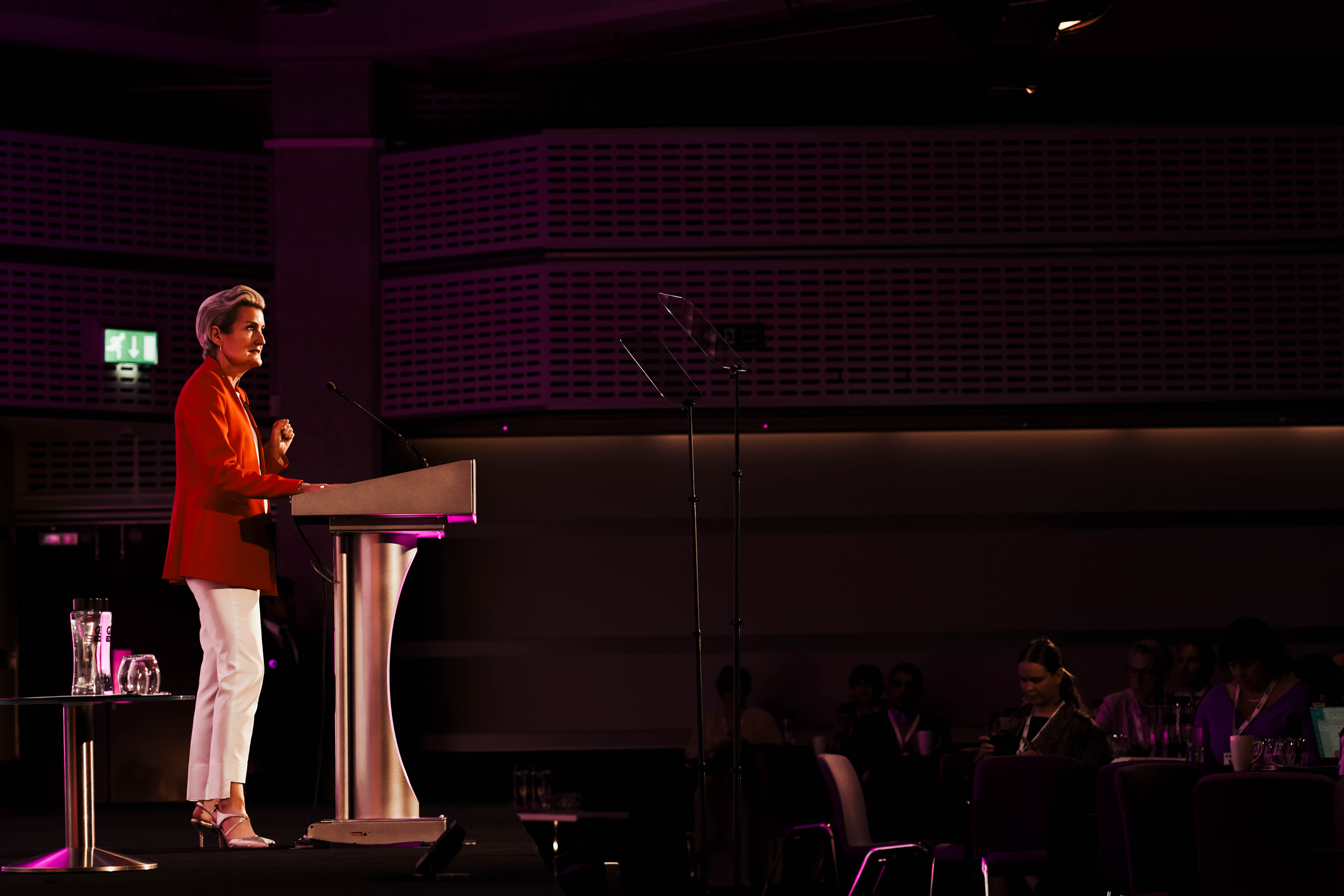 A woman with short blonde hair, dressed in a striking red blazer and white pants, stands at a podium and addresses an audience from a large, dimly lit stage with dramatic purple lighting.