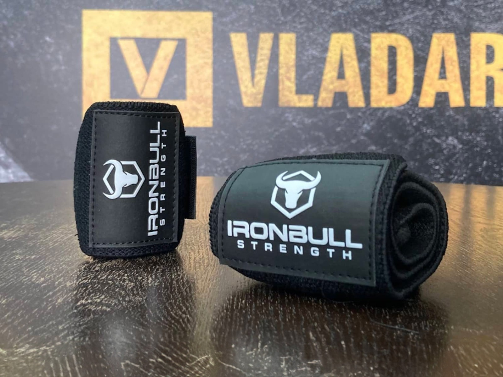 Black boxing/MMA hand wraps or gloves with "VLADAR" and "IRONBULL" branding displayed against a branded backdrop, showcasing sport equipment.