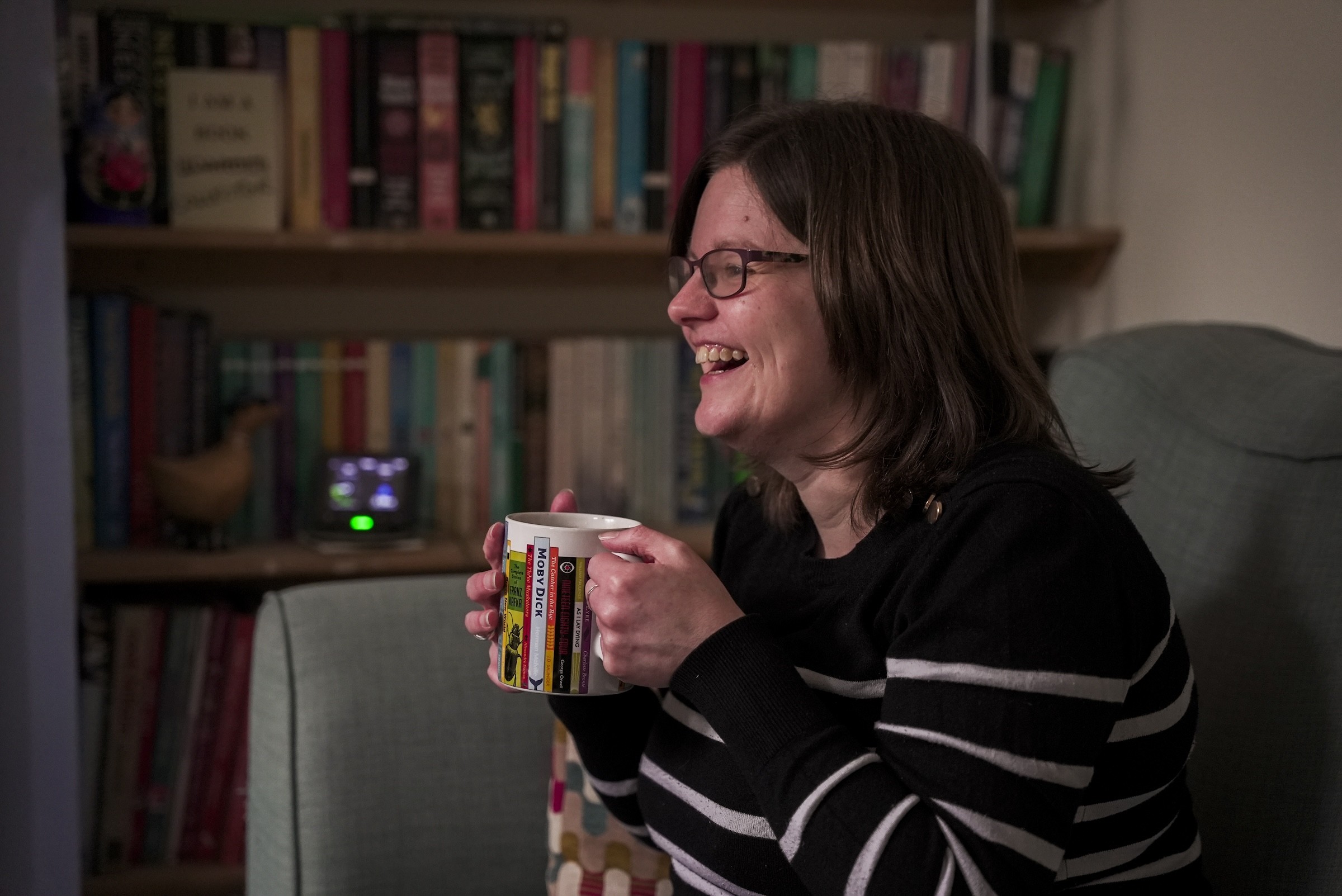 Woman sat on a sofa laughing and drinking a mug of tea.