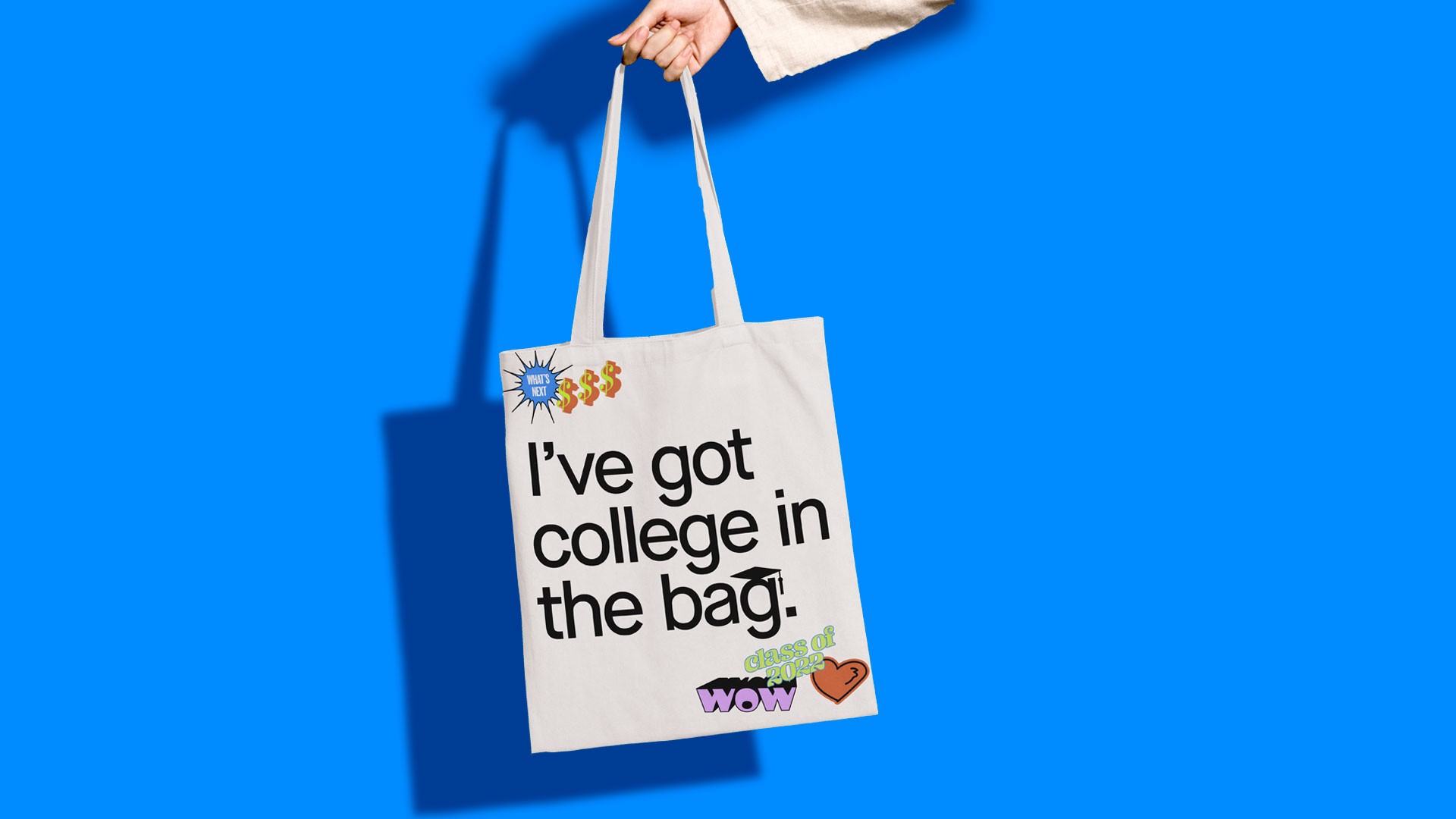 A hand holds a white tote bag with black text reading "I've got college in the bag." Small colorful stickers are affixed to the bag. The bag is set against a bright blue background.