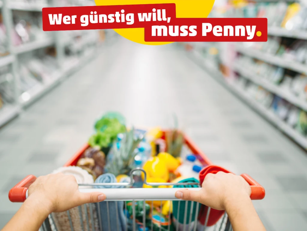 Eine Person schiebt einen vollen Einkaufswagen vor sich durch einen Gang in einem Einkaufsladen, auf dem Bild befinet sich der Claim der Firma Penny