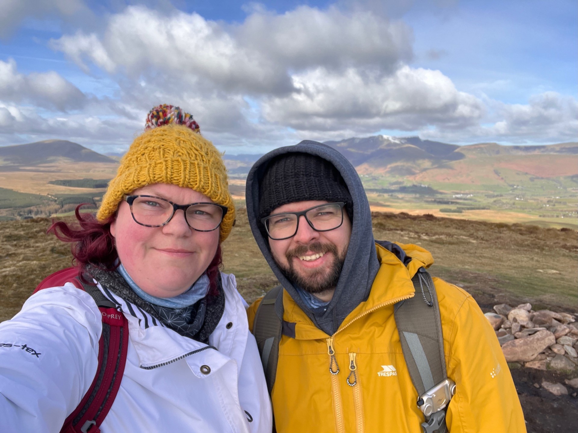 A selfie of Martin and April at the top of Great Mell Fell. Martin is wearing a yellow coat and a black hat, April is wearing a white coat and her yellow hat.