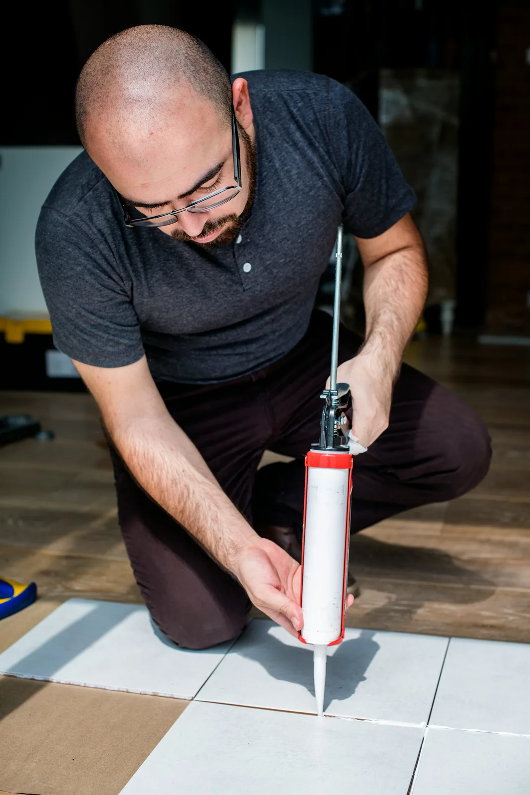 Man applying caulk sealant between white floor tiles with gun